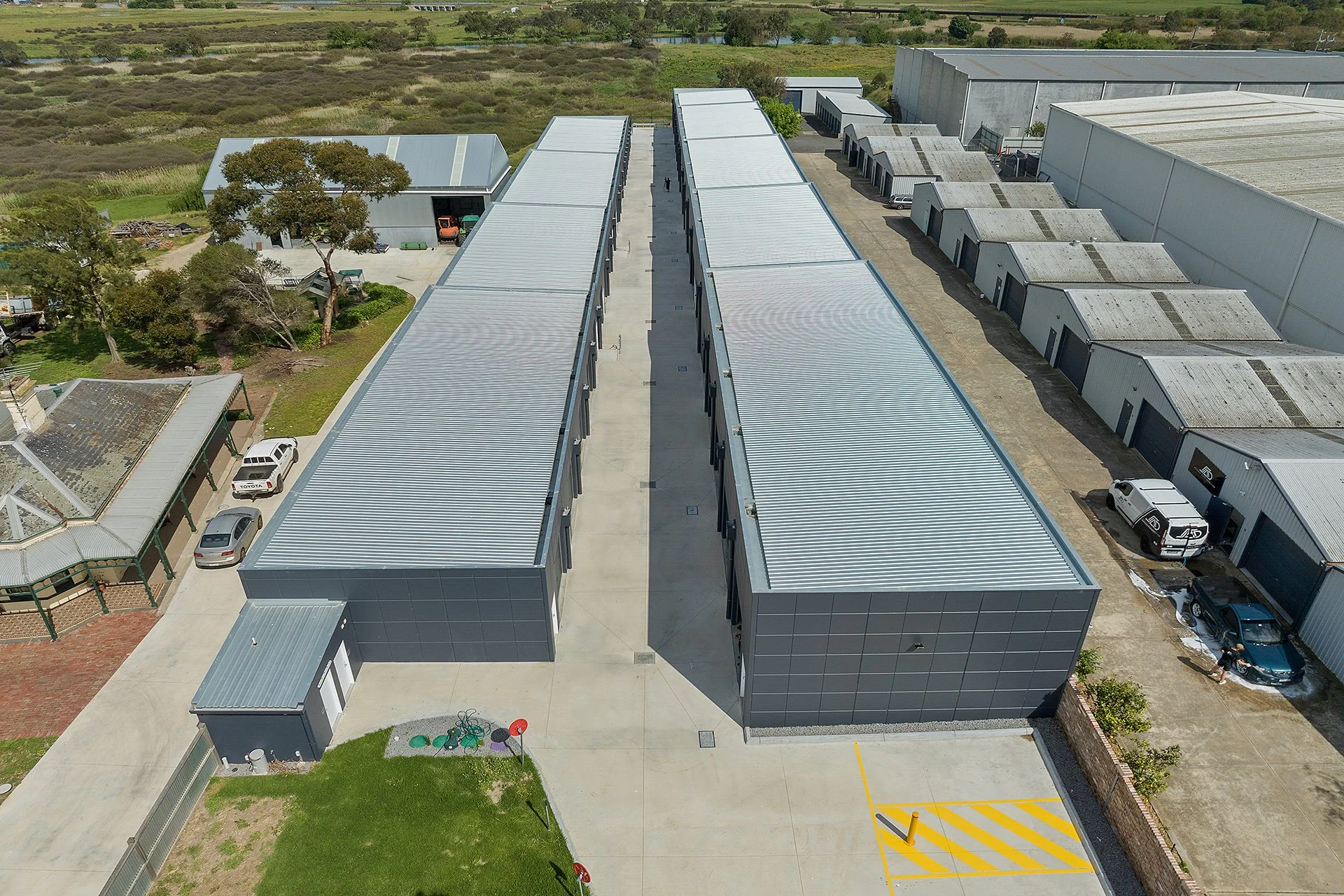 Aerial view of a modern industrial complex with multiple metal buildings, parked vehicles, and surrounding greenery.