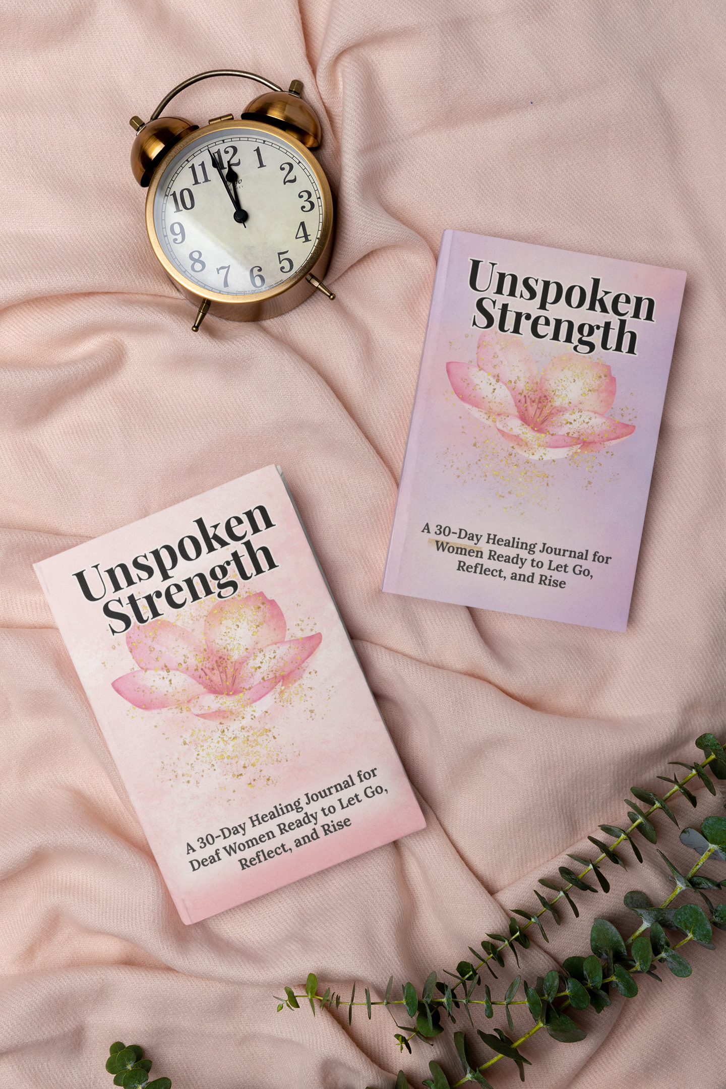 Two pink and purple books titled 'Unspoken Strength,' a gold vintage alarm clock showing 11:56, a sprig of green leaves, all on soft pink fabric.