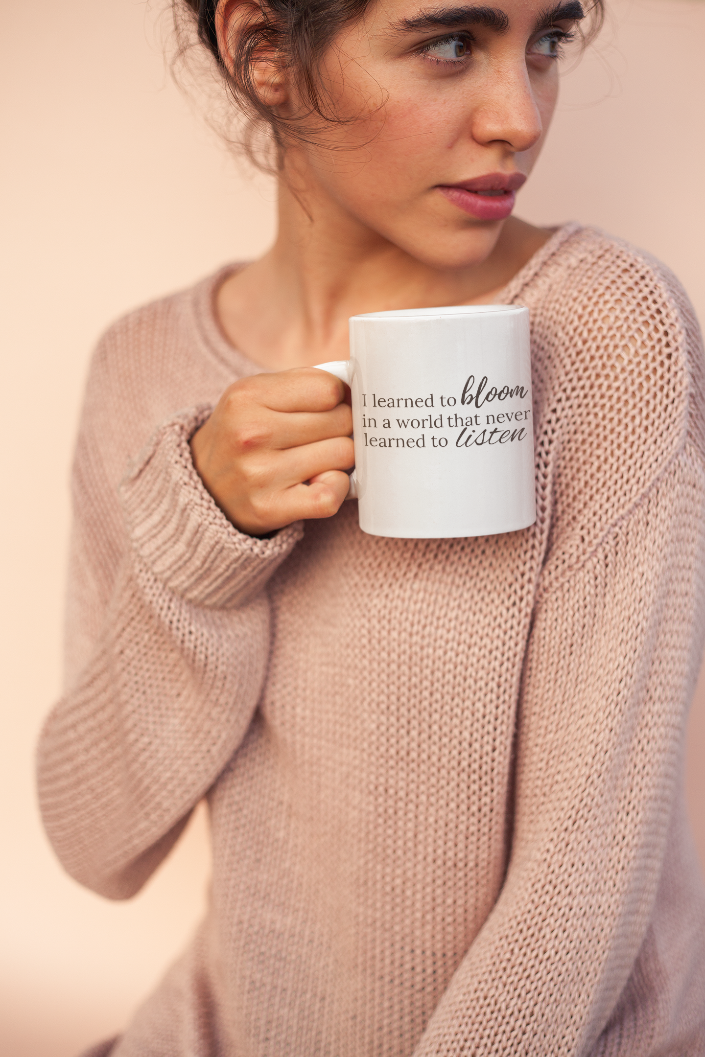 A woman with curly hair holding a white mug with a quote, wearing a pink knit sweater, against a soft peach background.