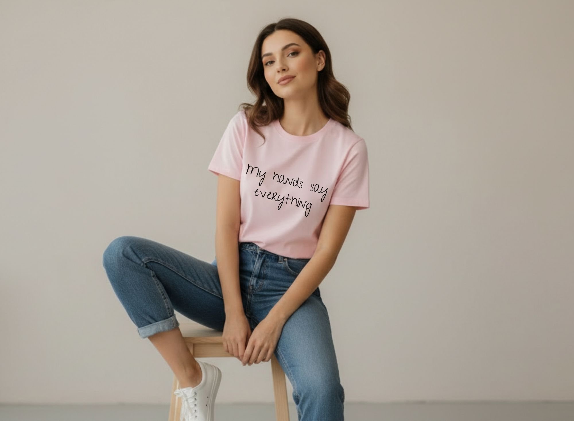 Woman wearing a pink “My Hands Say Everything” t-shirt, sitting on a wooden stool in a minimal room, representing silent expression and identity.