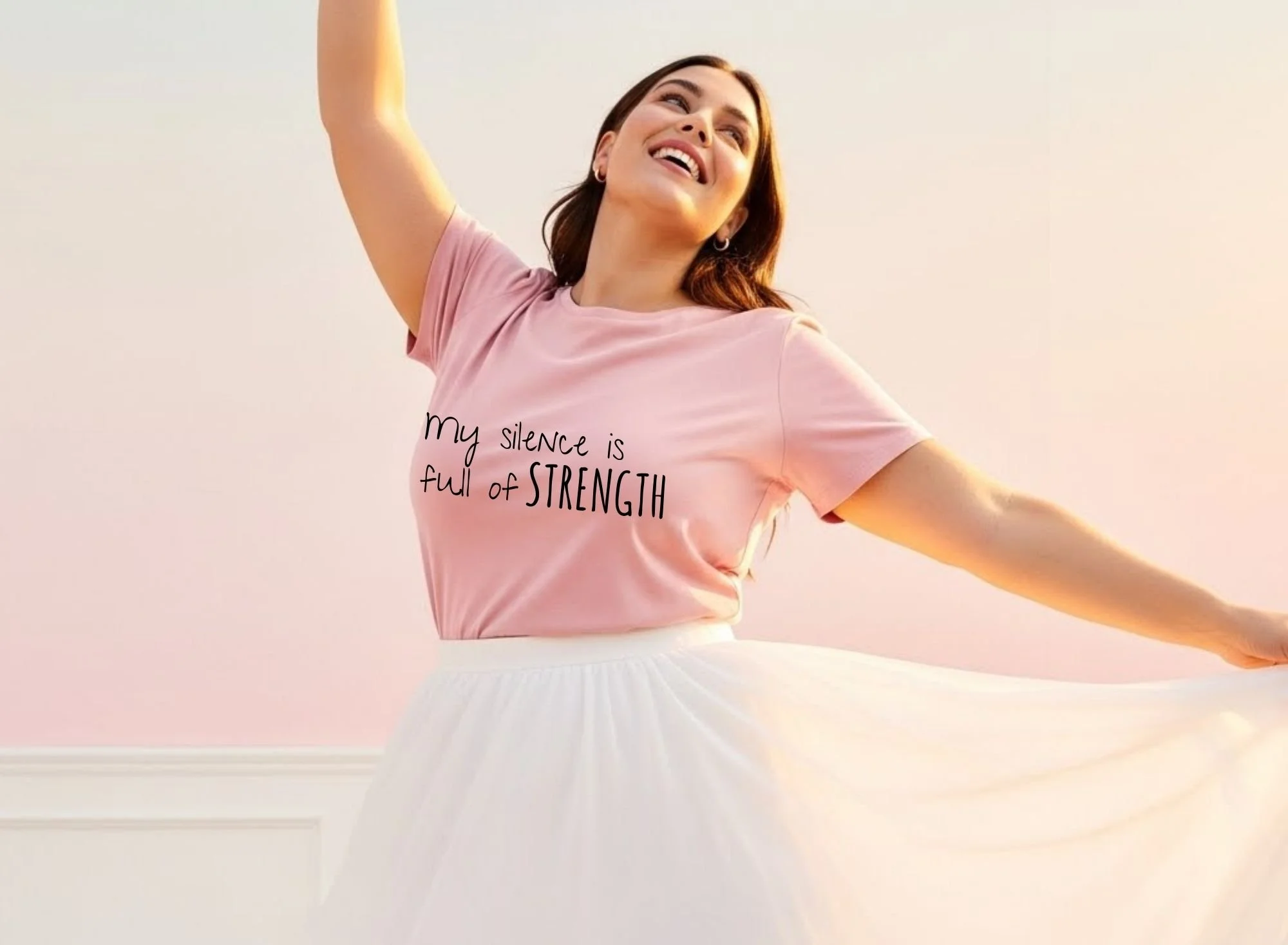 A woman smiling with arms outstretched, wearing a pink T-shirt that says 'my silence is full of STRENGTH' and a white skirt, standing against a light-colored wall.
