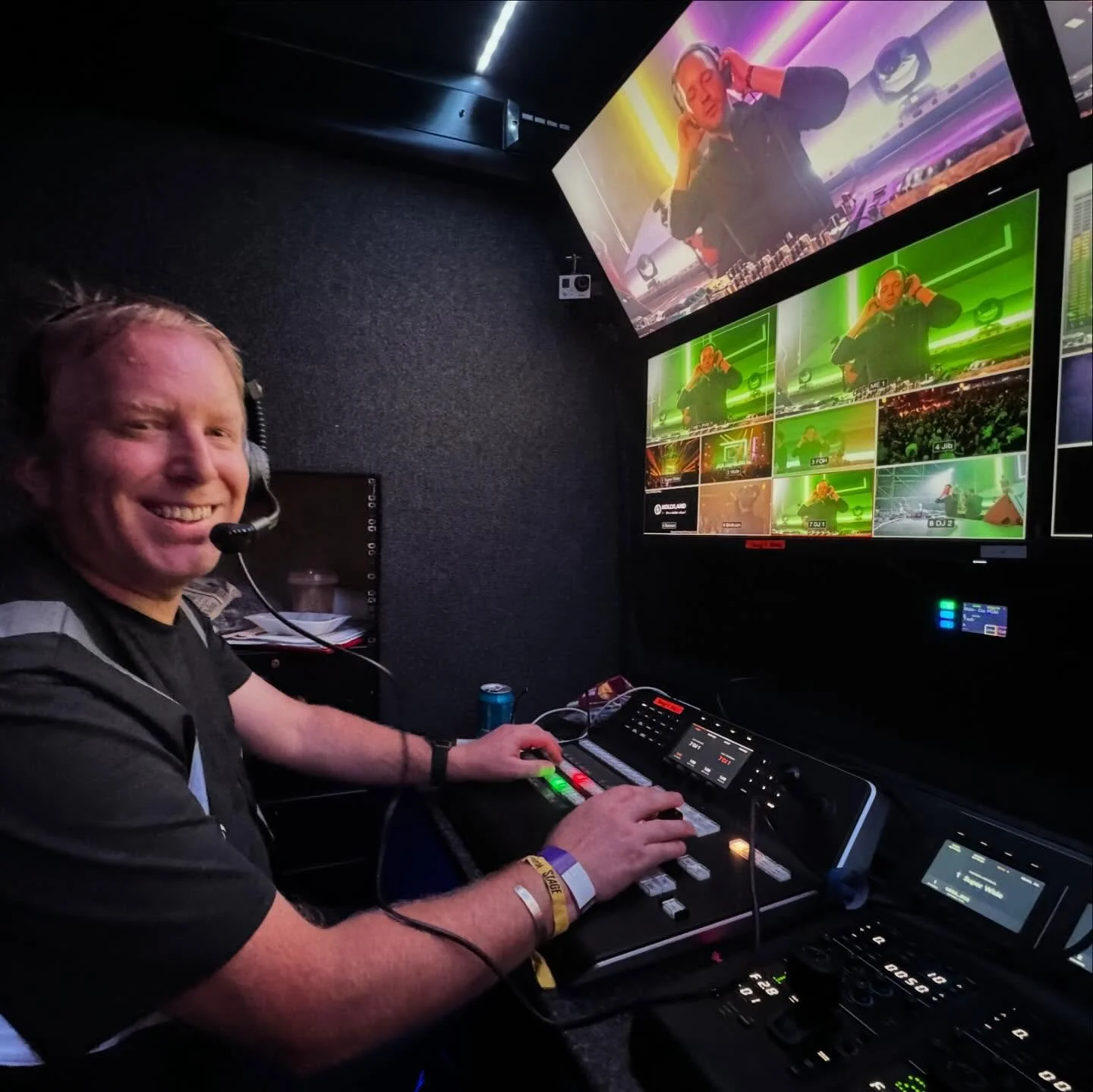 A smiling man wearing a headset and a wristband, operating a control panel inside a video studio or production room, with multiple monitors behind him displaying various scenes from a live event featuring a DJ with headphones in front of colorful lights.
