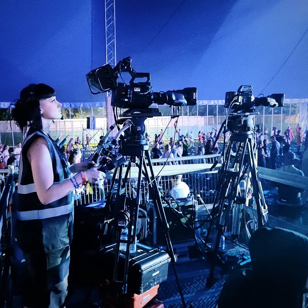 A woman operating a professional camera at an indoor event with a crowd in the background.