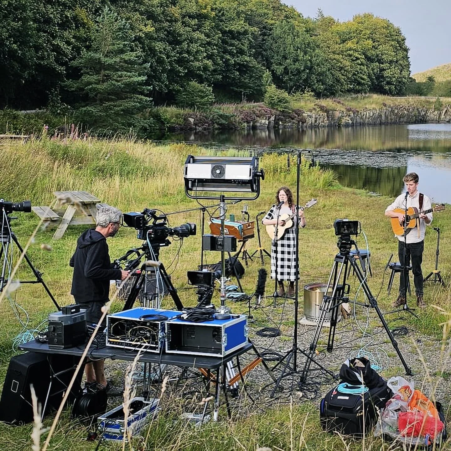 Musicians performing outdoors by a lake, with equipment and a camera crew filming.