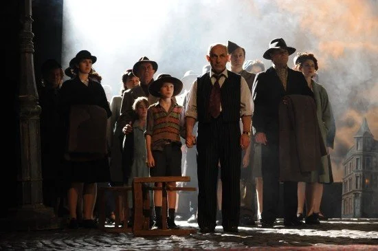 A group of people, including children and adults, dressed in early 20th-century clothing, standing on a street at night with smoke or fog in the background.