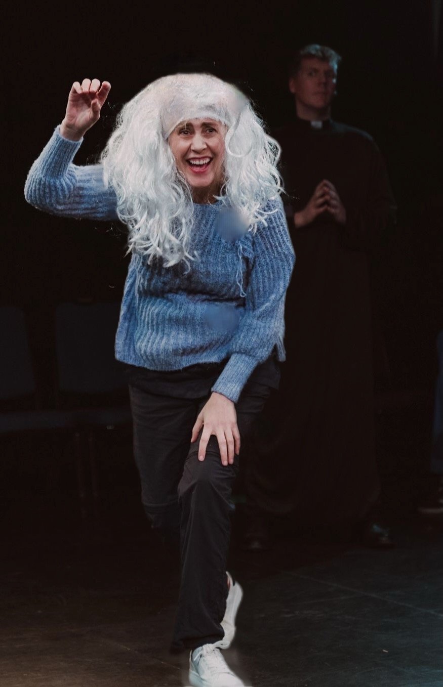 Woman with white wig dancing and smiling, wearing a blue sweater and black pants, in front of a man dressed as a priest in a dark room.