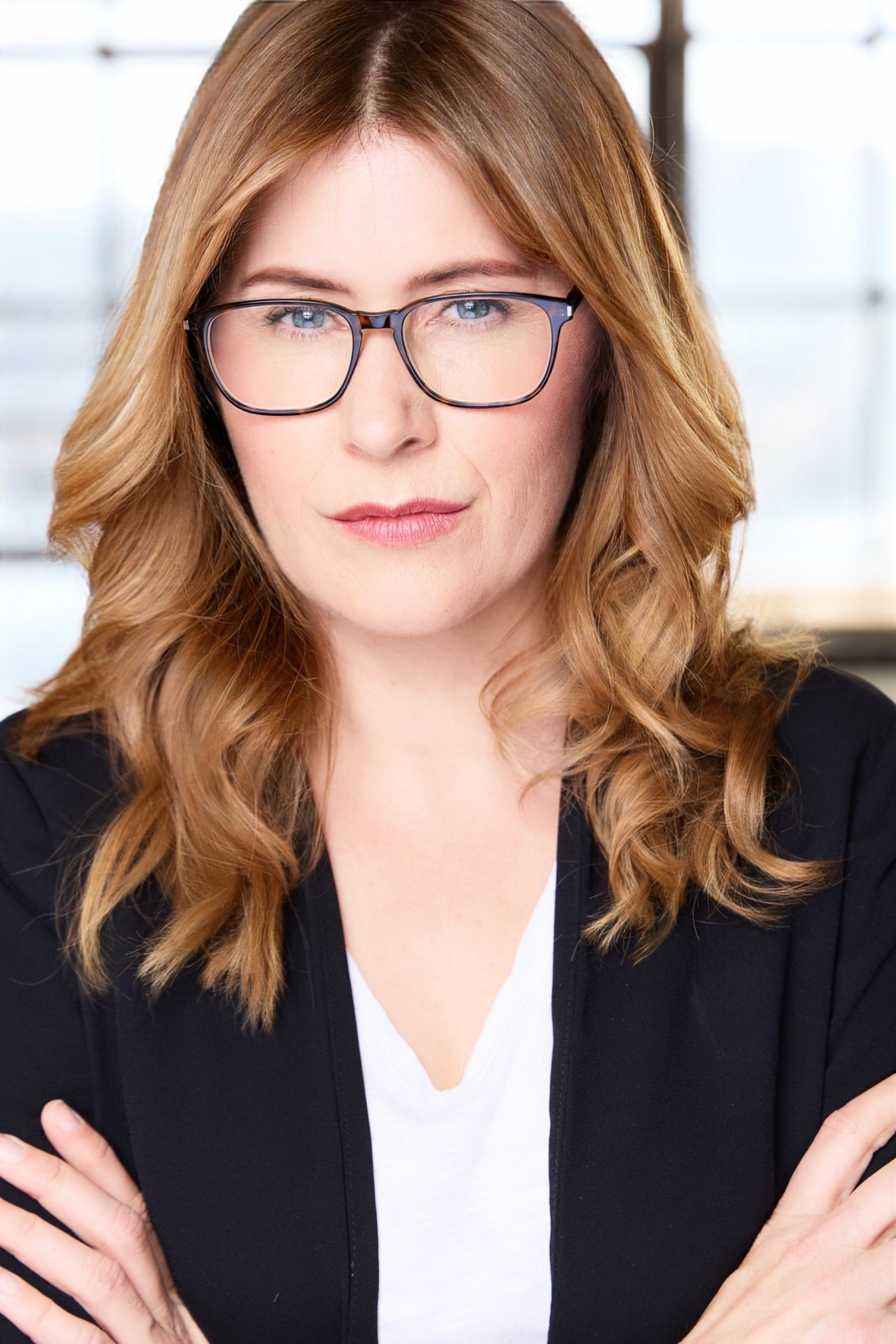 Close-up of a woman with long wavy red hair, glasses, wearing a black blazer and white shirt, crossing her arms.