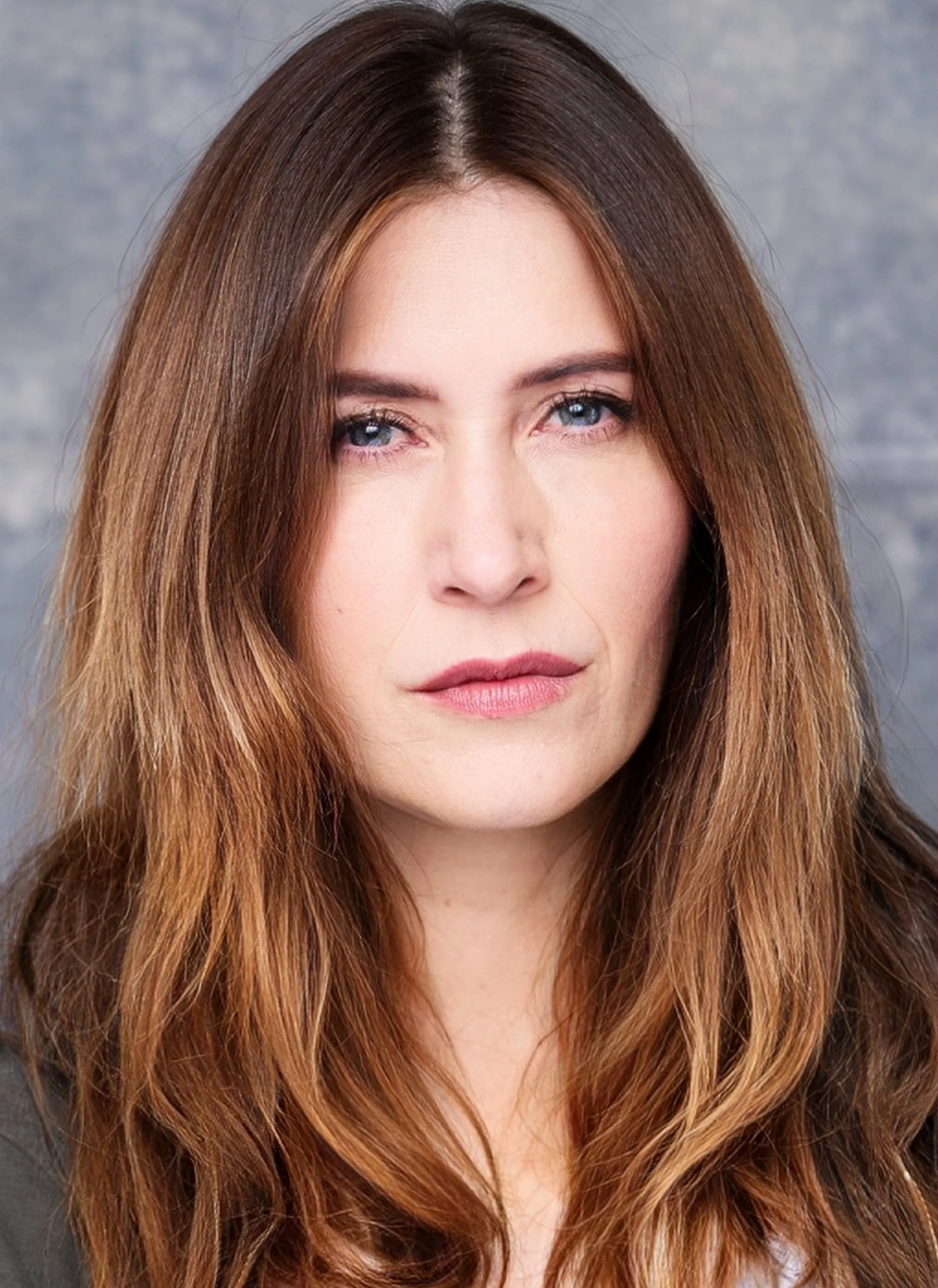 Close-up of a woman with long, wavy, reddish-brown hair and blue eyes, looking directly at the camera.