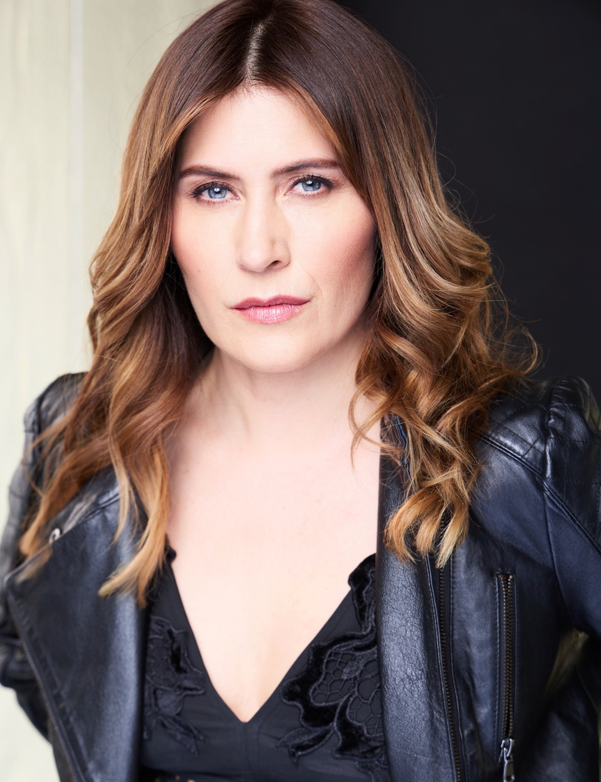 Close-up of a woman with long, wavy red hair, blue eyes, wearing natural makeup, a black lace top, and a black leather jacket with a zipper, against a black background.