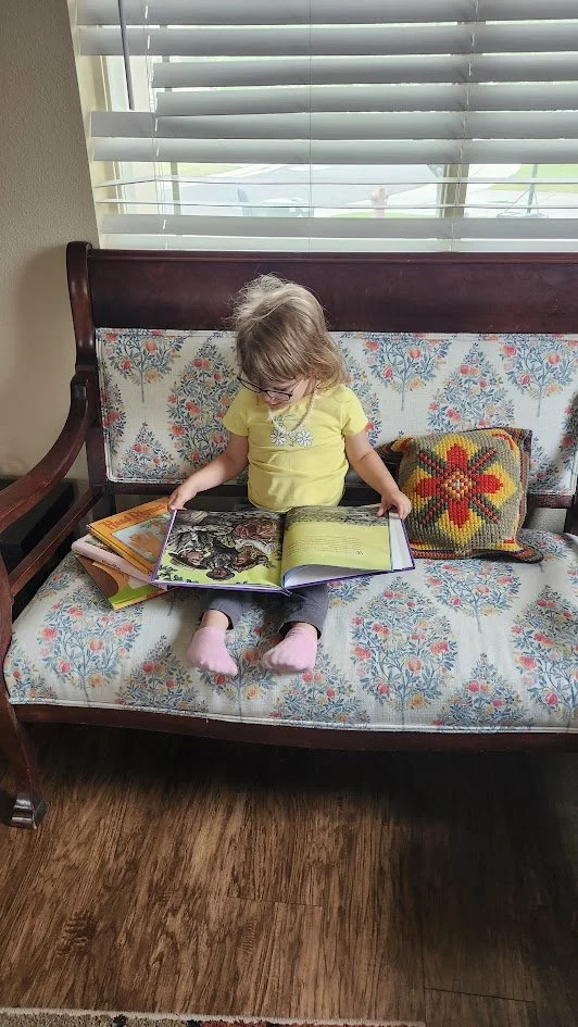 A small girl in a yellow t-shirt, pink socks, and pearl bead necklace reads a picture book
