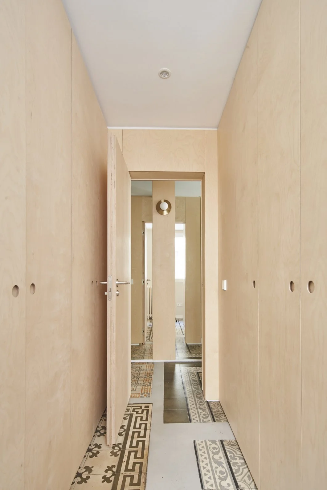 Pasillo con paredes de madera clara, piso de baldosas decorativas y puerta abierta con espejo en la pared.