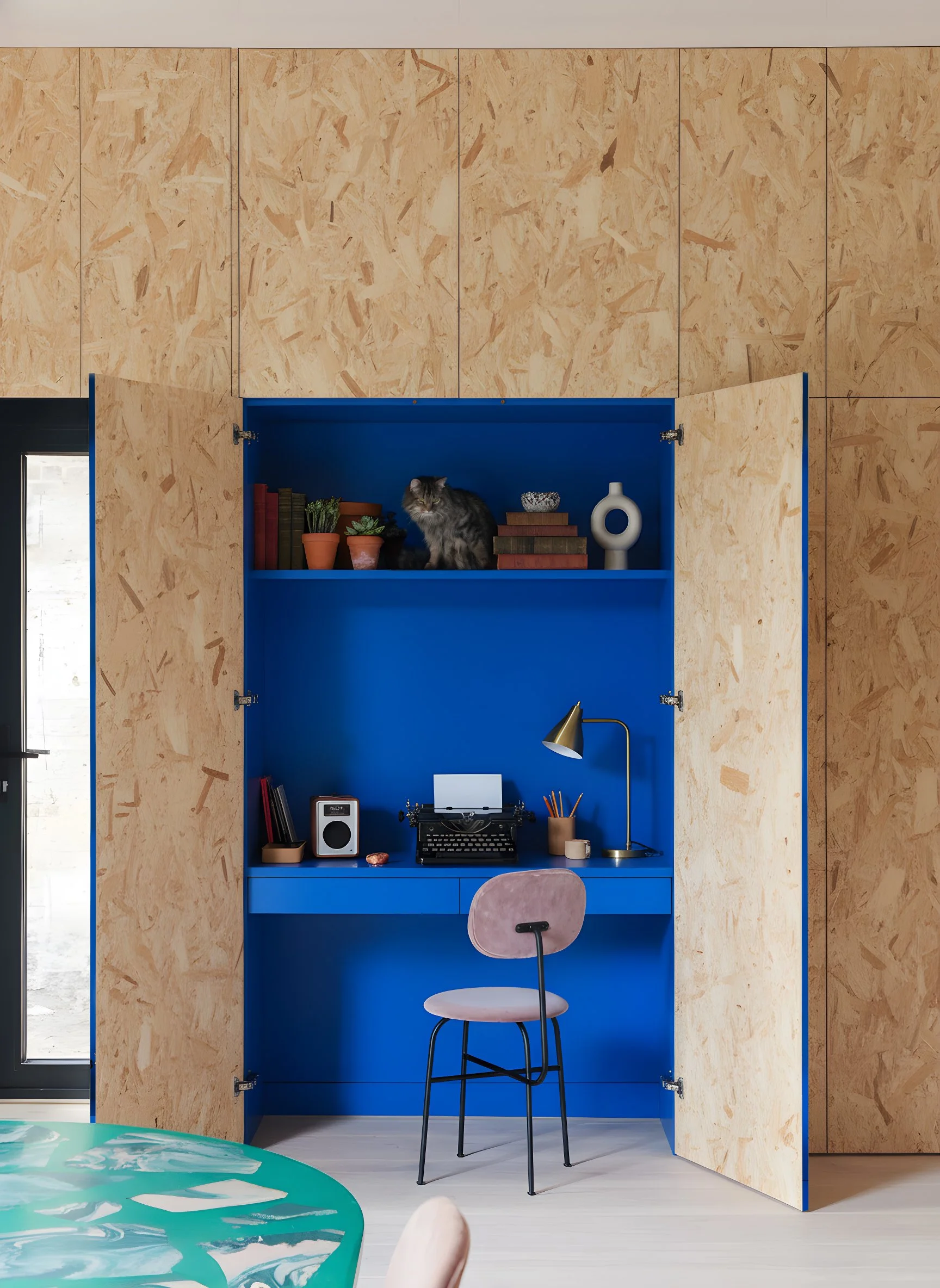 Estudio en un armario de madera con interior azul, con una sala y una silla rosa, objetos decorativos y un gato en la estantería.