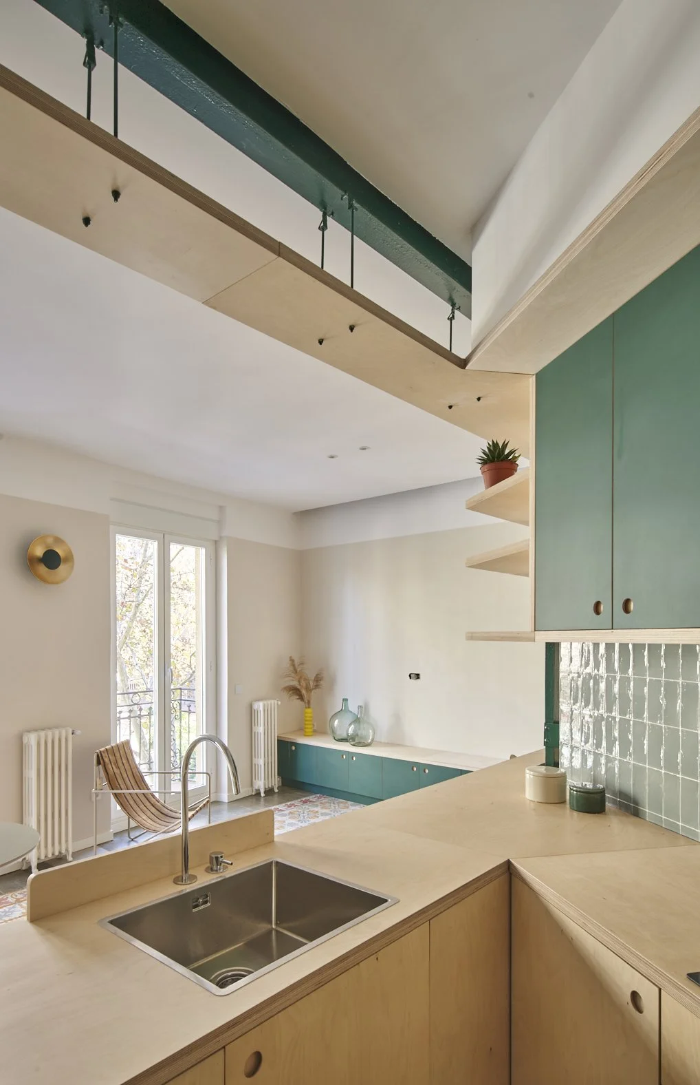 Vista de una sala y cocina moderna con cocina de barra de madera clara, gabinetes verdes, decoraciones con plantas y botellas, y un balcón con puertas de cristal.