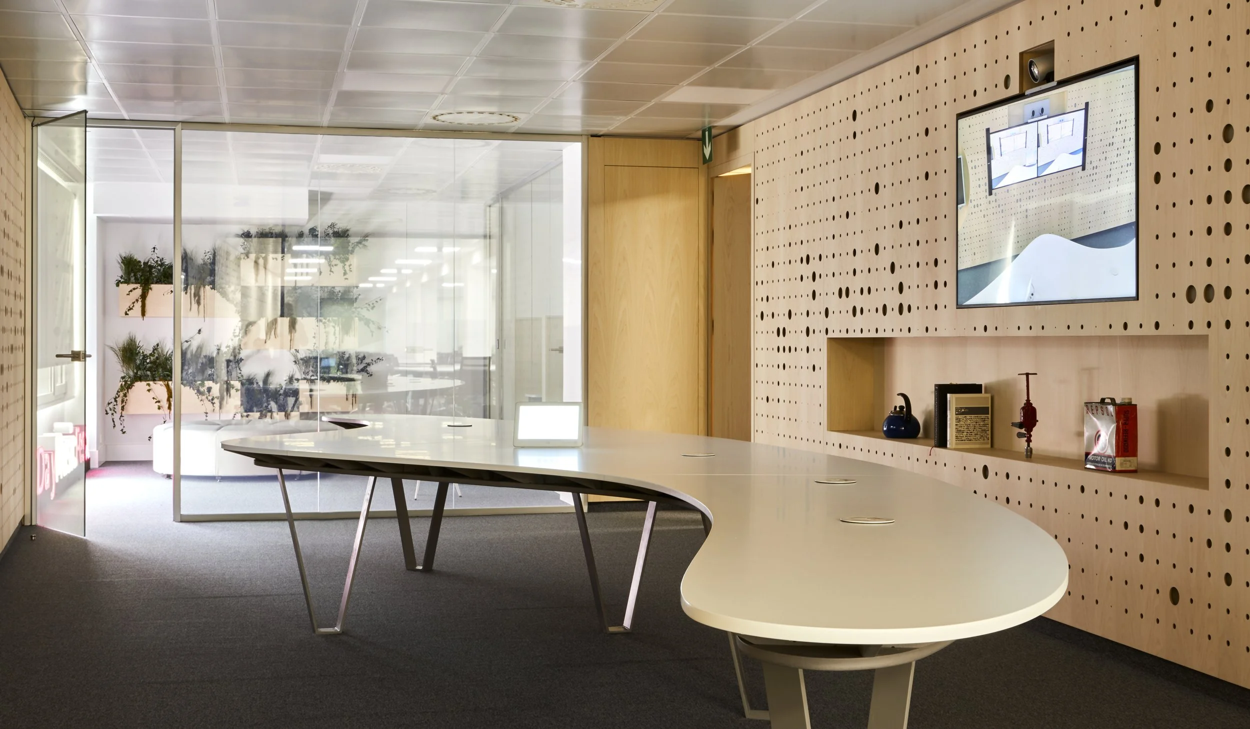 Sala de reuniones con mesa curva y pared de paneles perforados, pantalla y estantería con objetos decorativos y alimentos.
