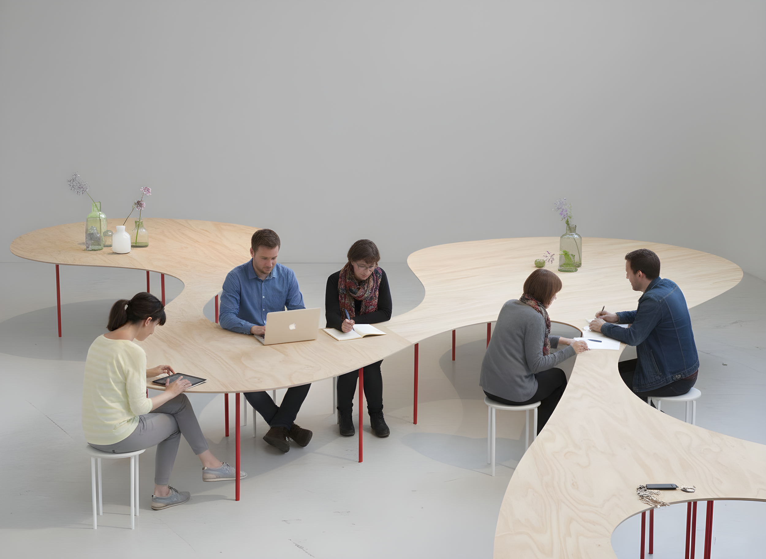 Grupo de seis personas sentadas en mesas de madera curvadas en un espacio minimalista, con vasos con flores en el fondo.