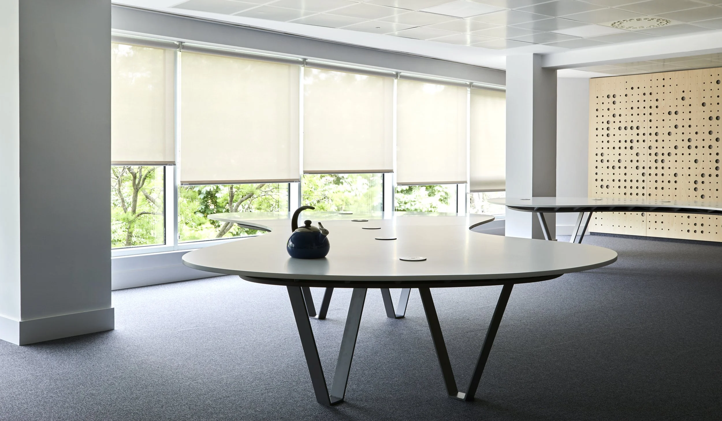 Sala de reuniones moderna con una mesa redonda blanca y una tetera azul en el centro, ventanas grandes con persianas blancas y pared de madera perforada.