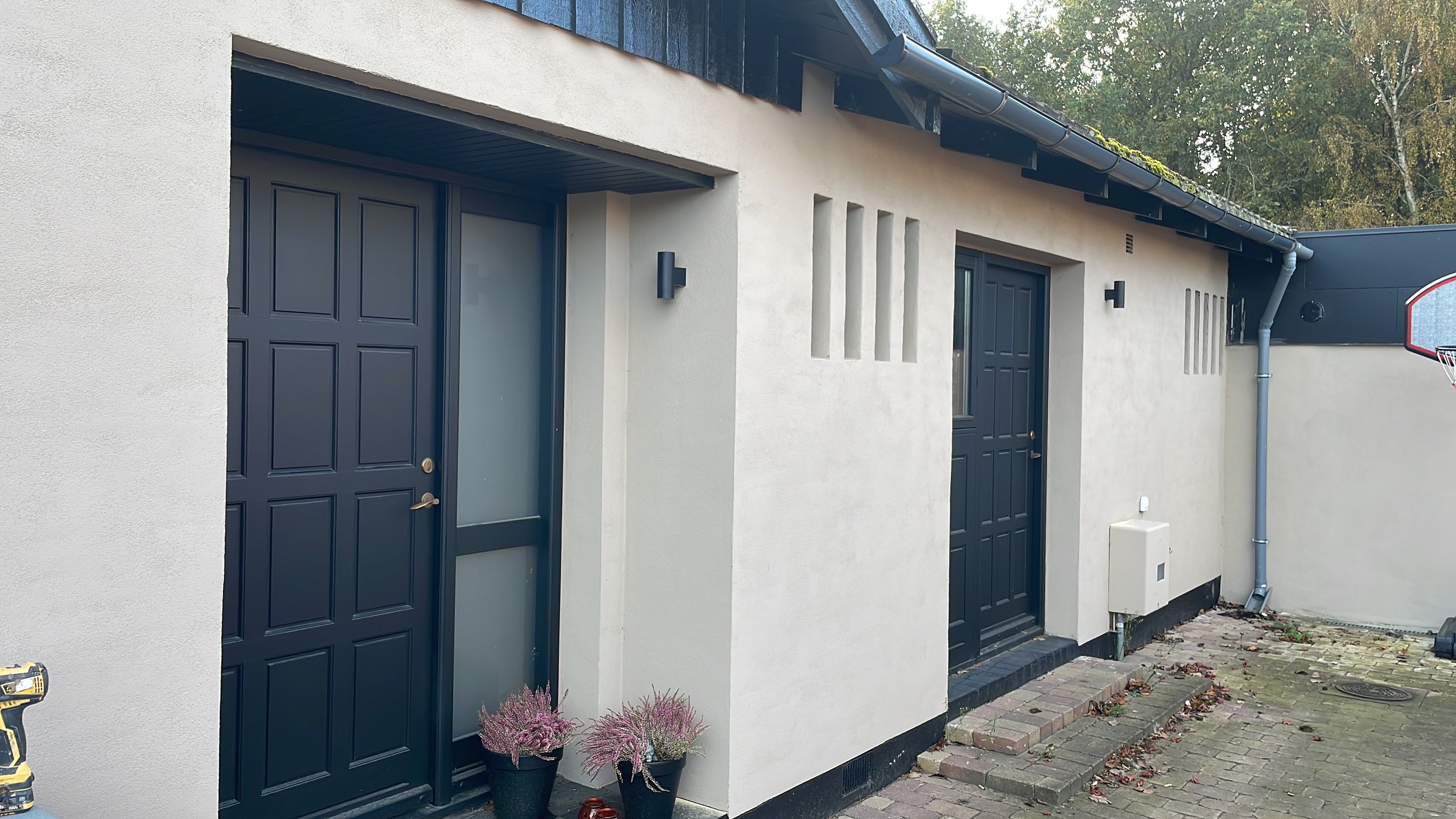 Exterior view of a modern house with two black doors, one with a glass panel, and a window in between. Two black wall-mounted lights are on the wall, and there are two potted plants with pinkish flowers on the ground near the left door. The house has a light-colored stucco finish with dark trim and a paved walkway in front.