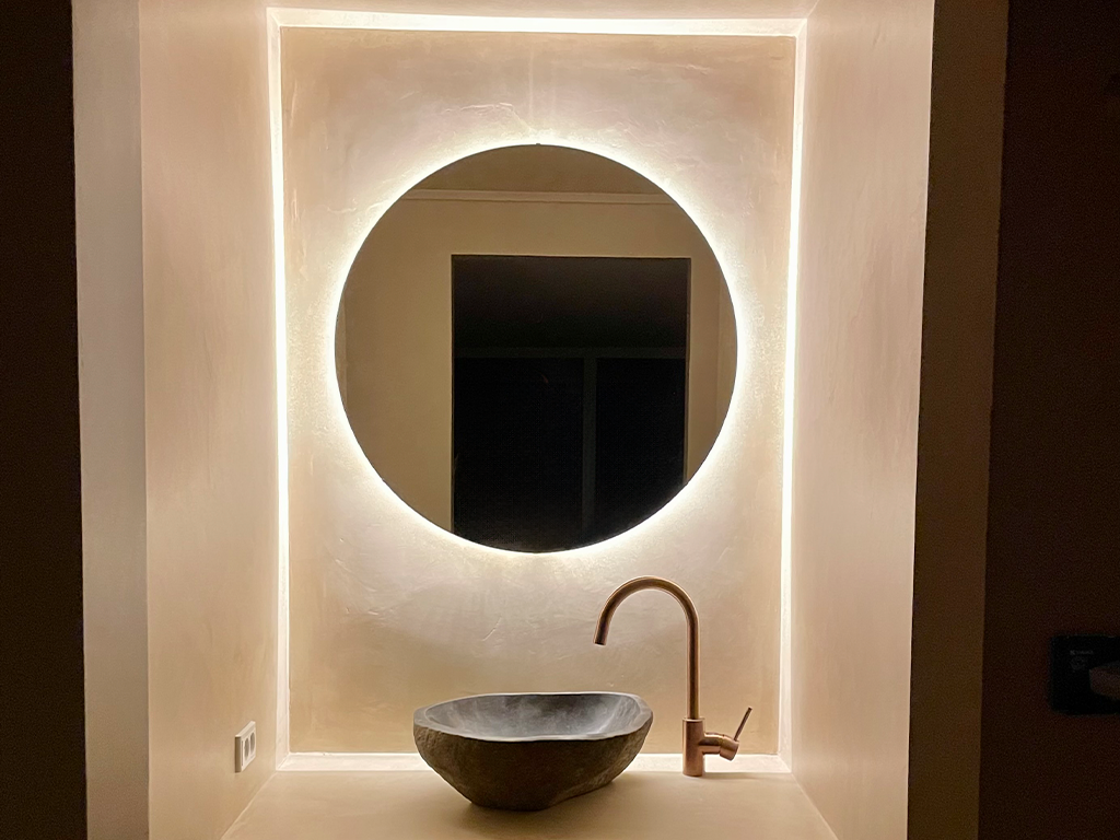 A modern bathroom with a backlit round mirror, a stone vessel sink, and a gold faucet.