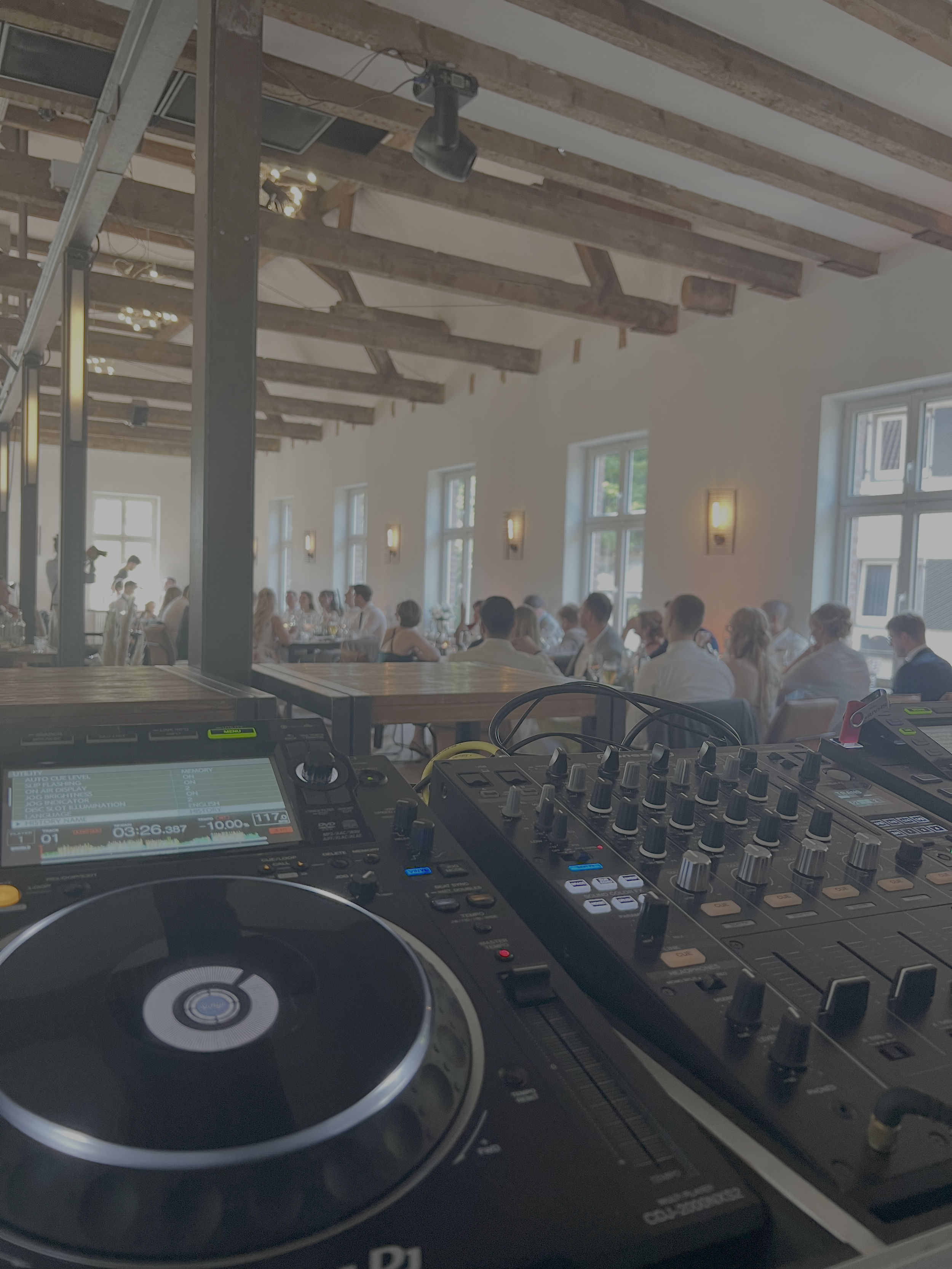 Blick auf eine DJ-Konsole in einem Veranstaltungsraum mit mehreren Gästen, die am Tisch sitzen, helle Wände, große Fenster und sichtbare Holzbalkendecke.