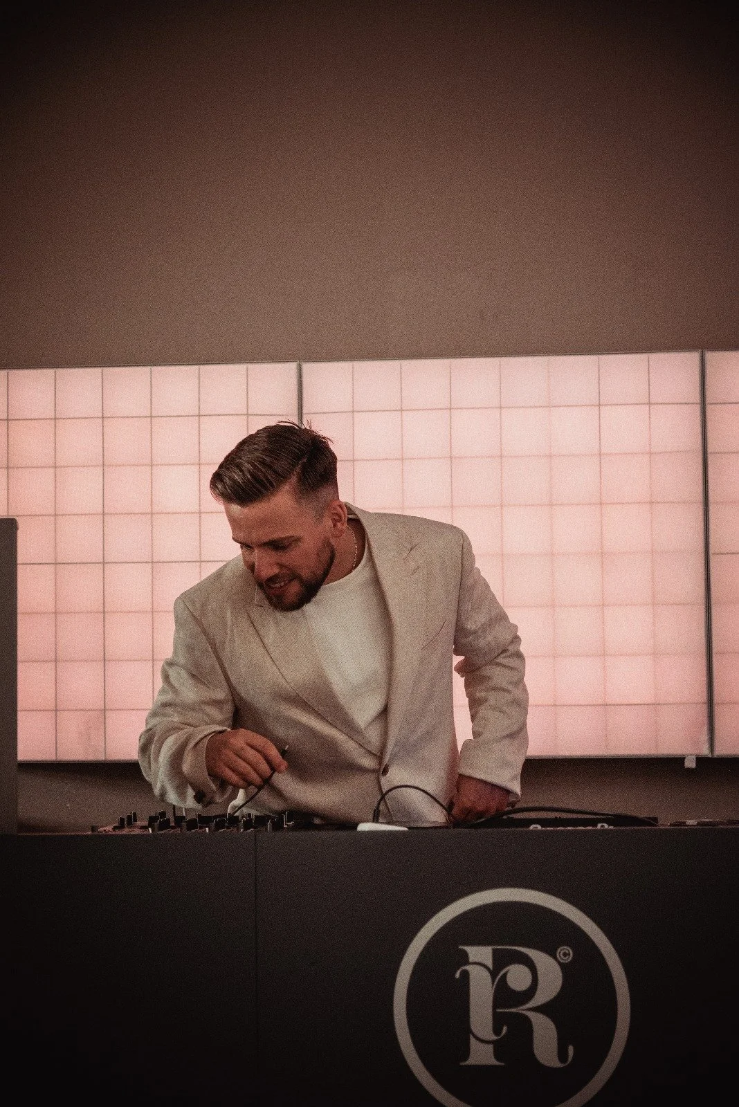 Ein Mann mit einem cremefarbenen Anzug und weißem Hemd, der am DJ-Pult auflegt und lächelt, im Hintergrund eine große, hell beleuchtete Wand.