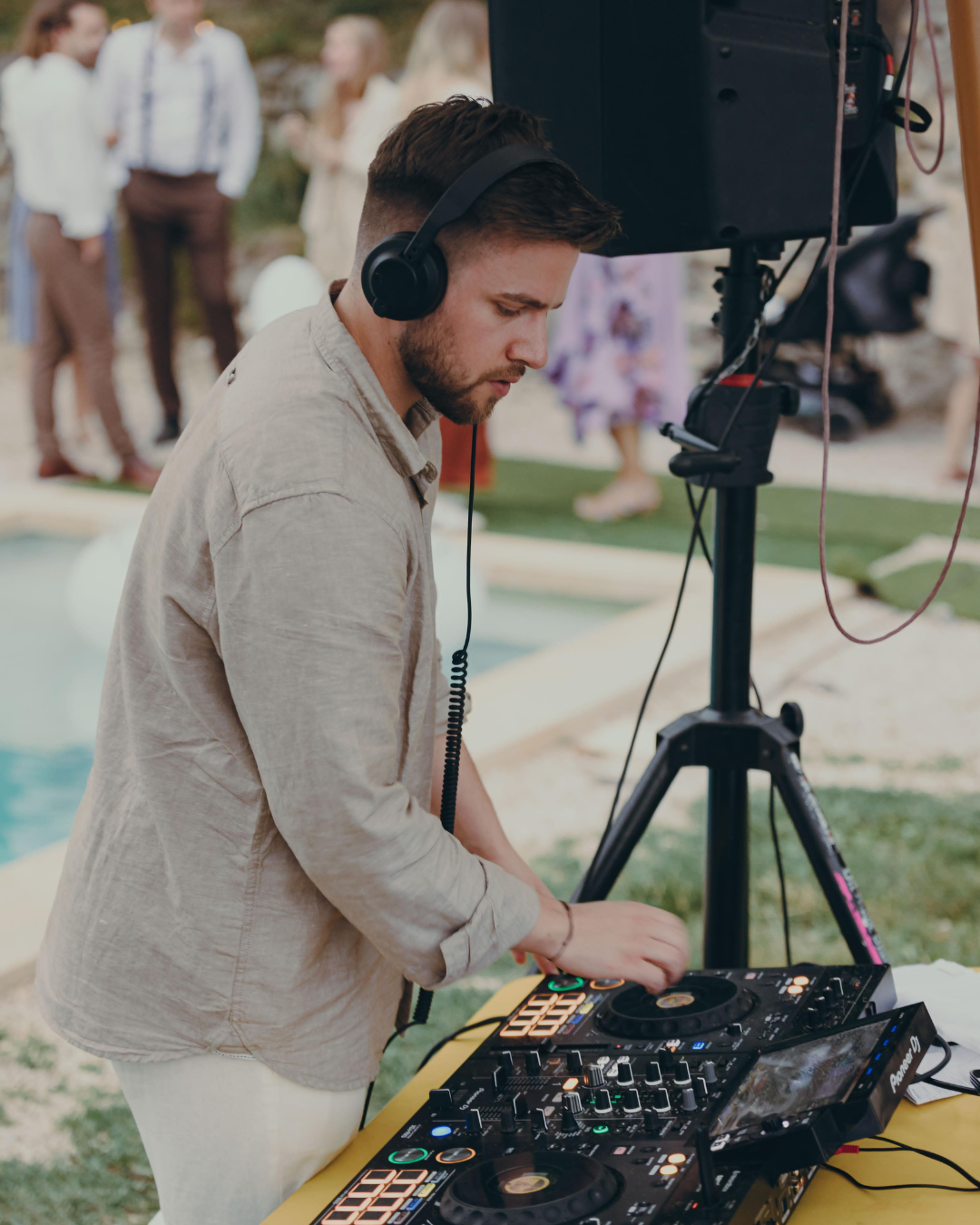 Ein Mann mit Kopfhörern bedient DJ-Equipment an einem Outdoor-Event, im Hintergrund sind mehrere Personen, die sich unterhalten und Zweige sichtbar.