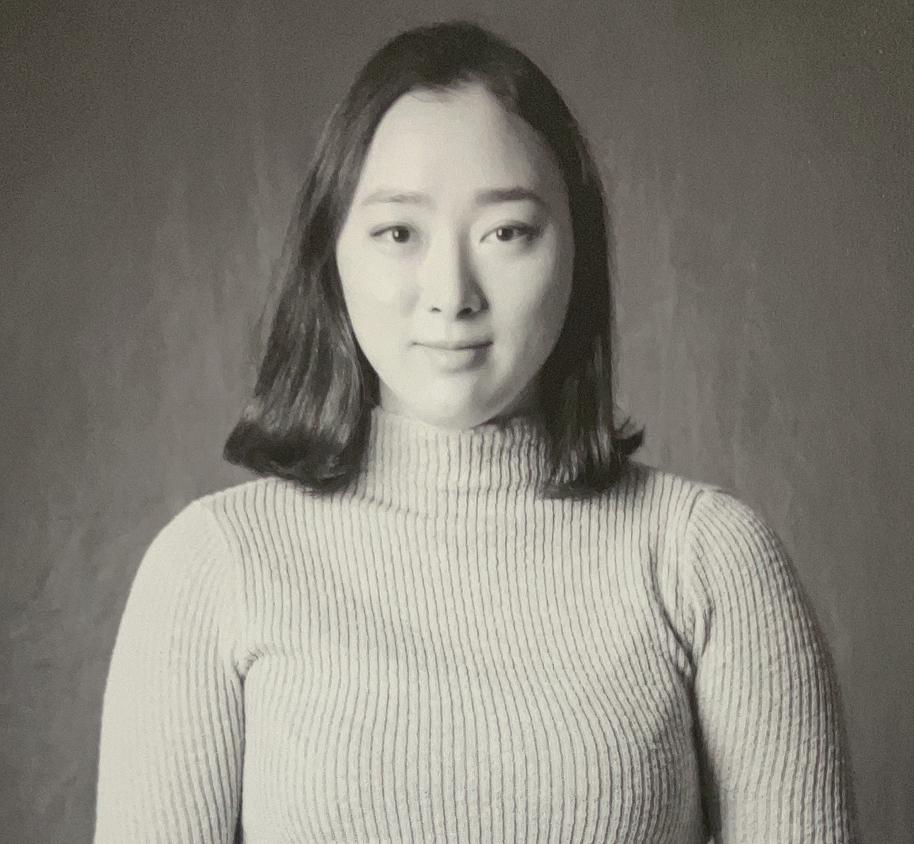 Black and white portrait of a young Asian woman with shoulder-length hair, wearing a ribbed turtleneck sweater, standing against a plain background.