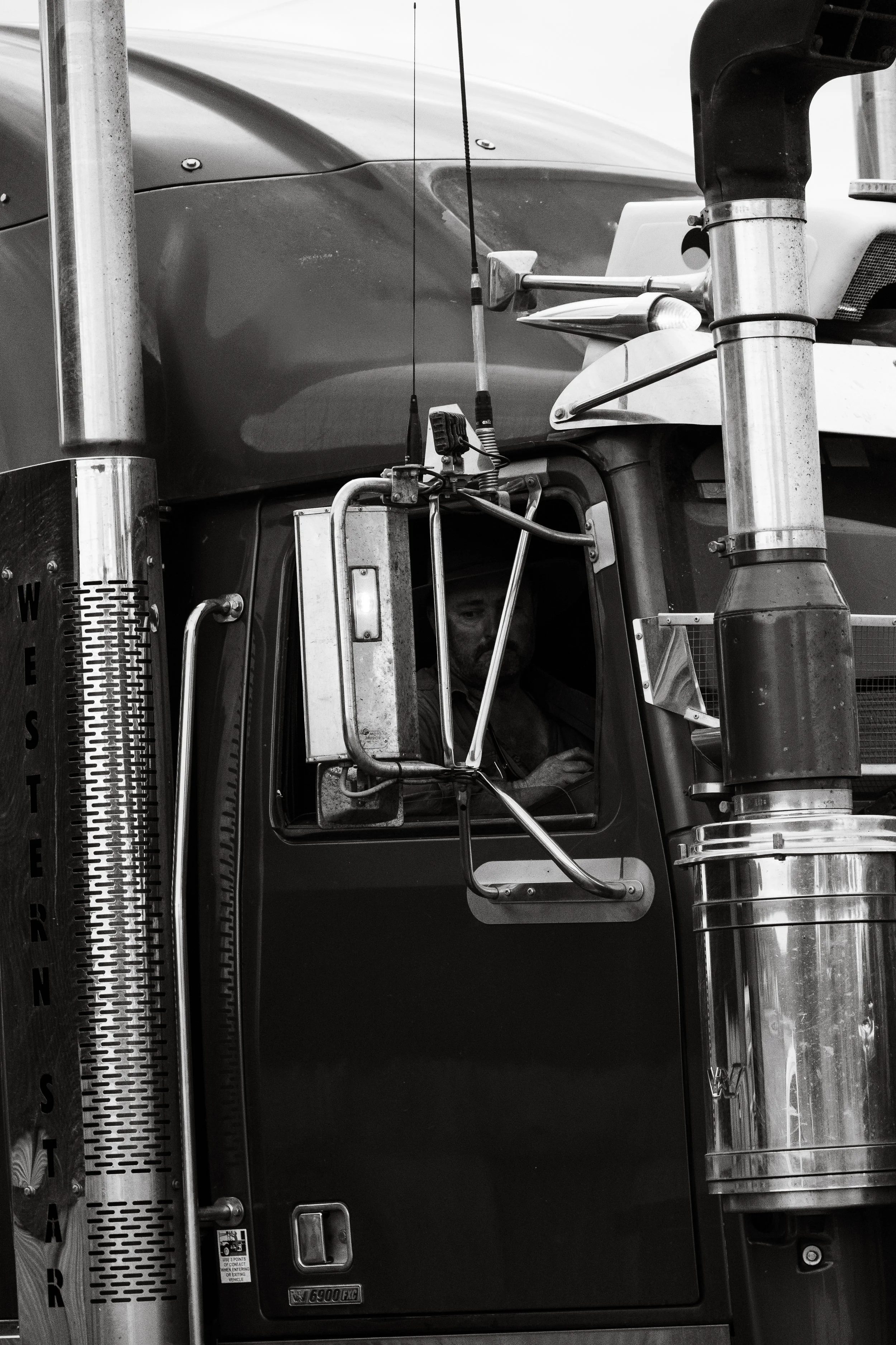 Black and white photo of a semi-truck's cab, focusing on the driver's side window where a man is sitting inside, looking out. The front of the truck features visible exhaust pipes, antennas, and metallic components.