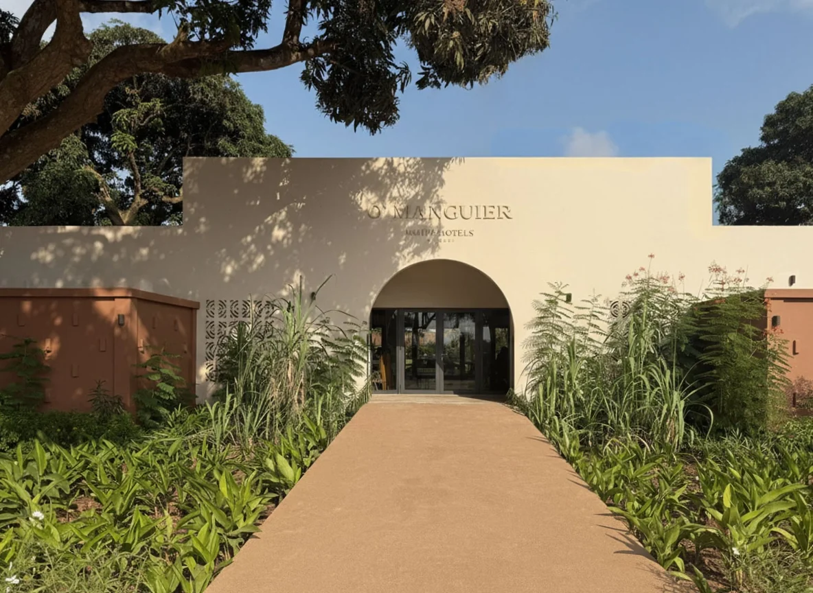 Ingresso di un hotel con un sentiero di fronte e piante verdi ai lati, sotto un cielo blu con alcune nuvole.