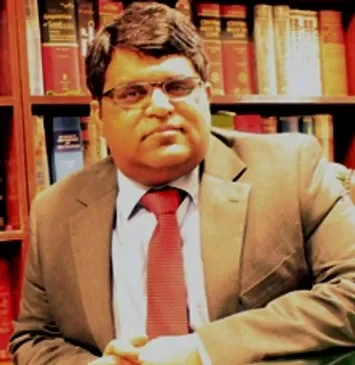 A man wearing glasses in a beige suit with a red tie, sitting in front of a bookshelf filled with books.