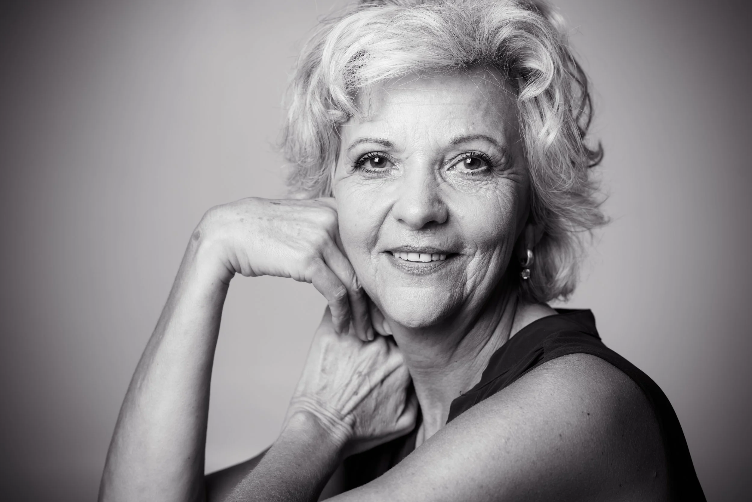 Schwarz-weiß Porträt einer älteren Frau mit lockigem Haar, die lächelnd in die Kamera schaut, mit ihrer Hand am Hals, elegant gekleidet und mit einem Ohrring.