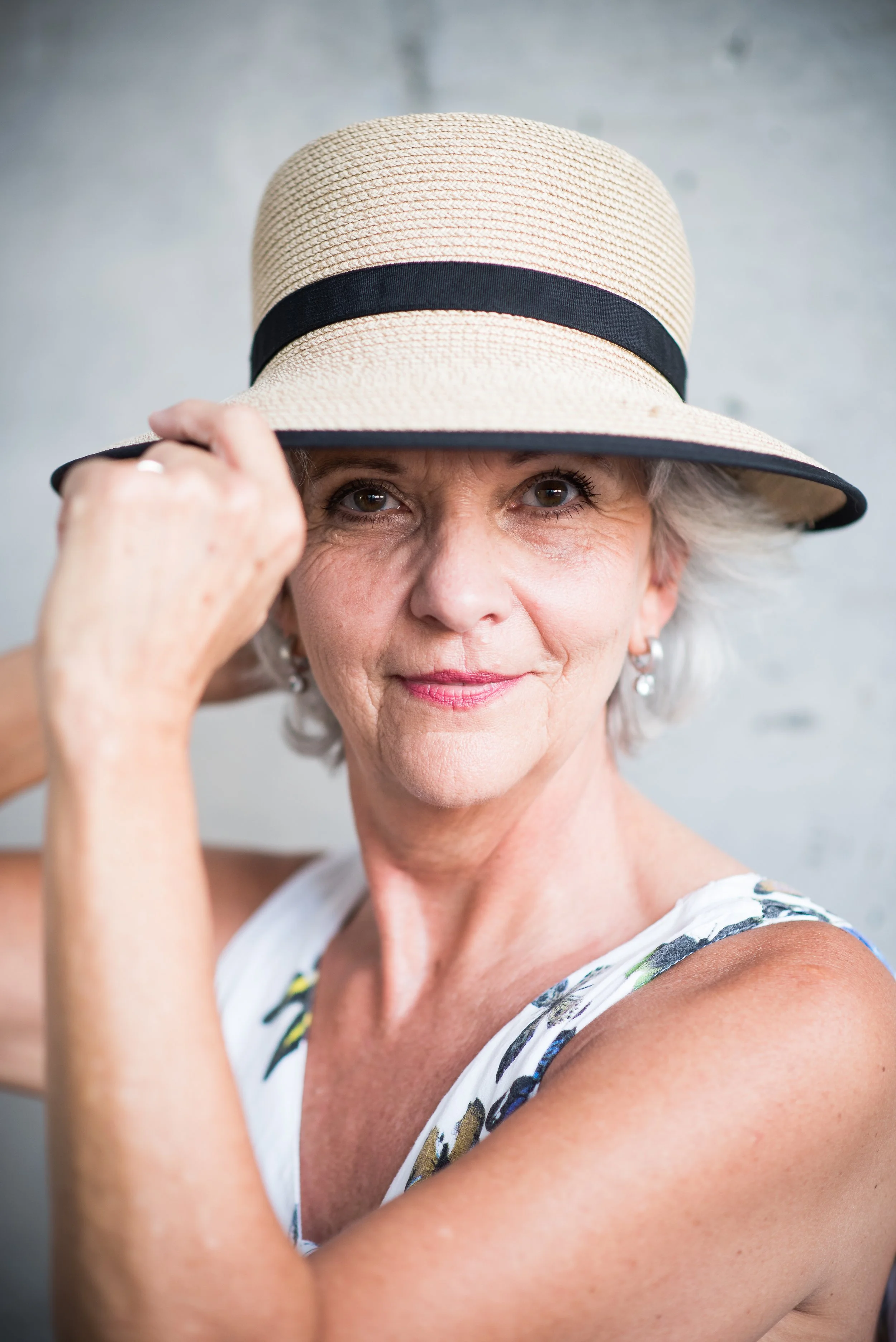 Eine ältere Frau trägt einen Strohhut mit schwarzem Band und ein sommerliches, ärmelloses Kleid mit Blumenmuster. Sie schaut in die Kamera und hebt den Hut leicht an, während sie ein leichtes Lächeln zeigt.
