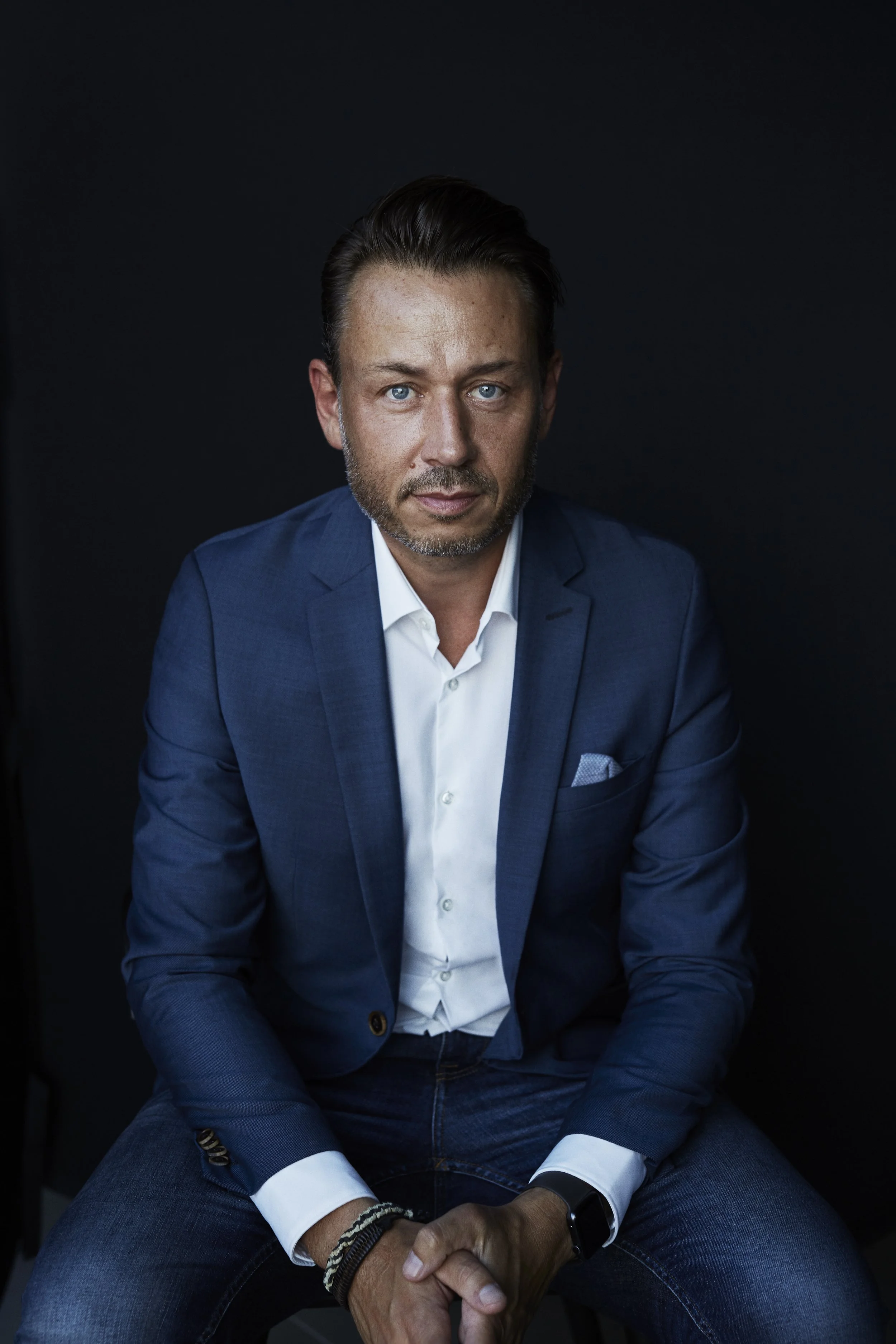A man in a blue suit and white shirt sitting against a dark background.