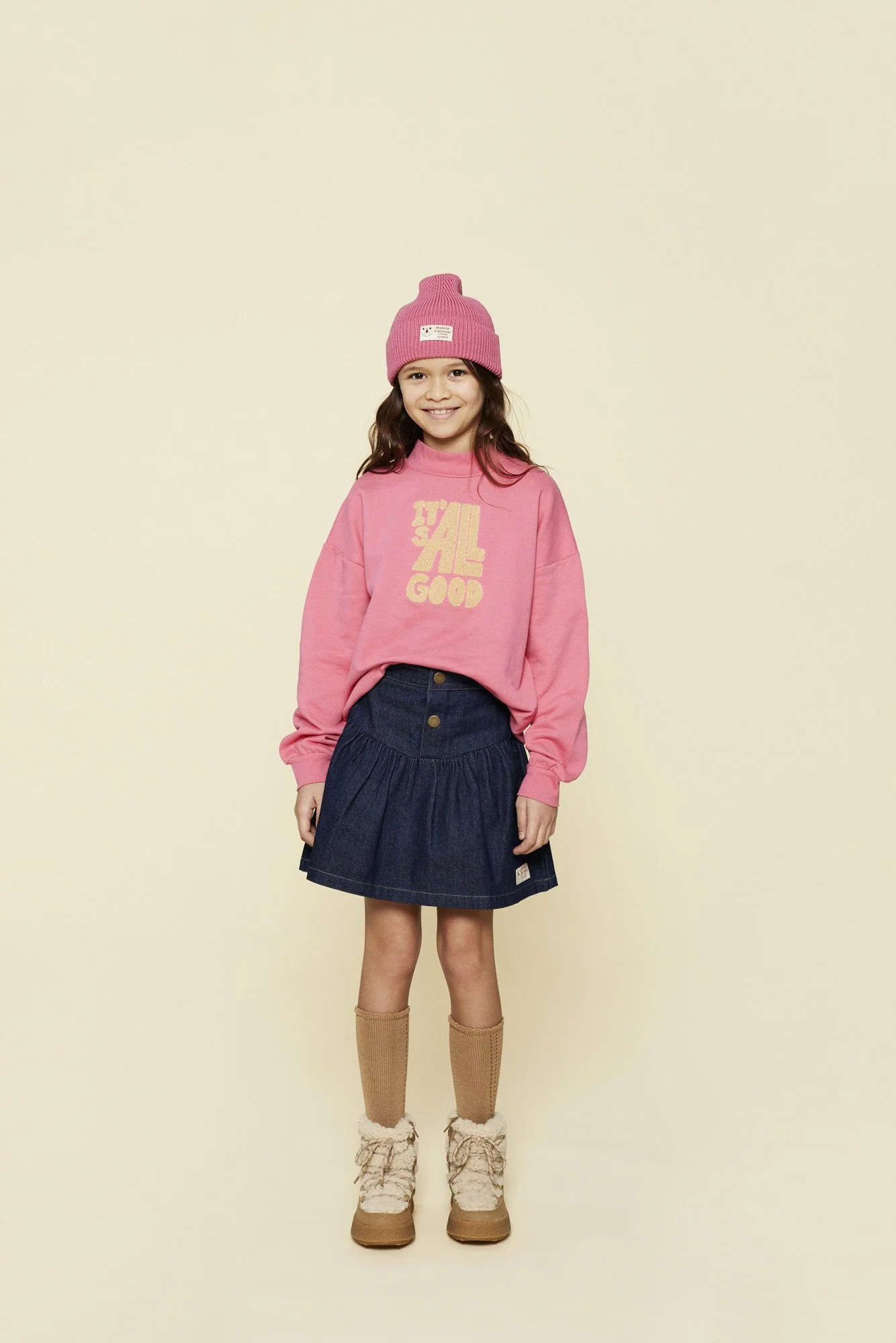 A young girl wearing a pink beanie hat, a pink sweatshirt with yellow text that reads 'IT'S ALL GOOD', a dark blue pleated skirt with buttons, tan knee-high socks, and tan boots with lace trim, smiling against a plain light background.
