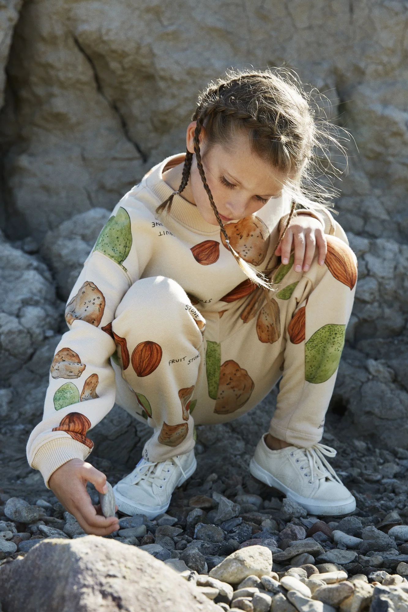 Child crouching outdoors on rocky terrain, wearing a cream-colored sweatshirt and pants with a nut and fruit print, holding a small stone.
