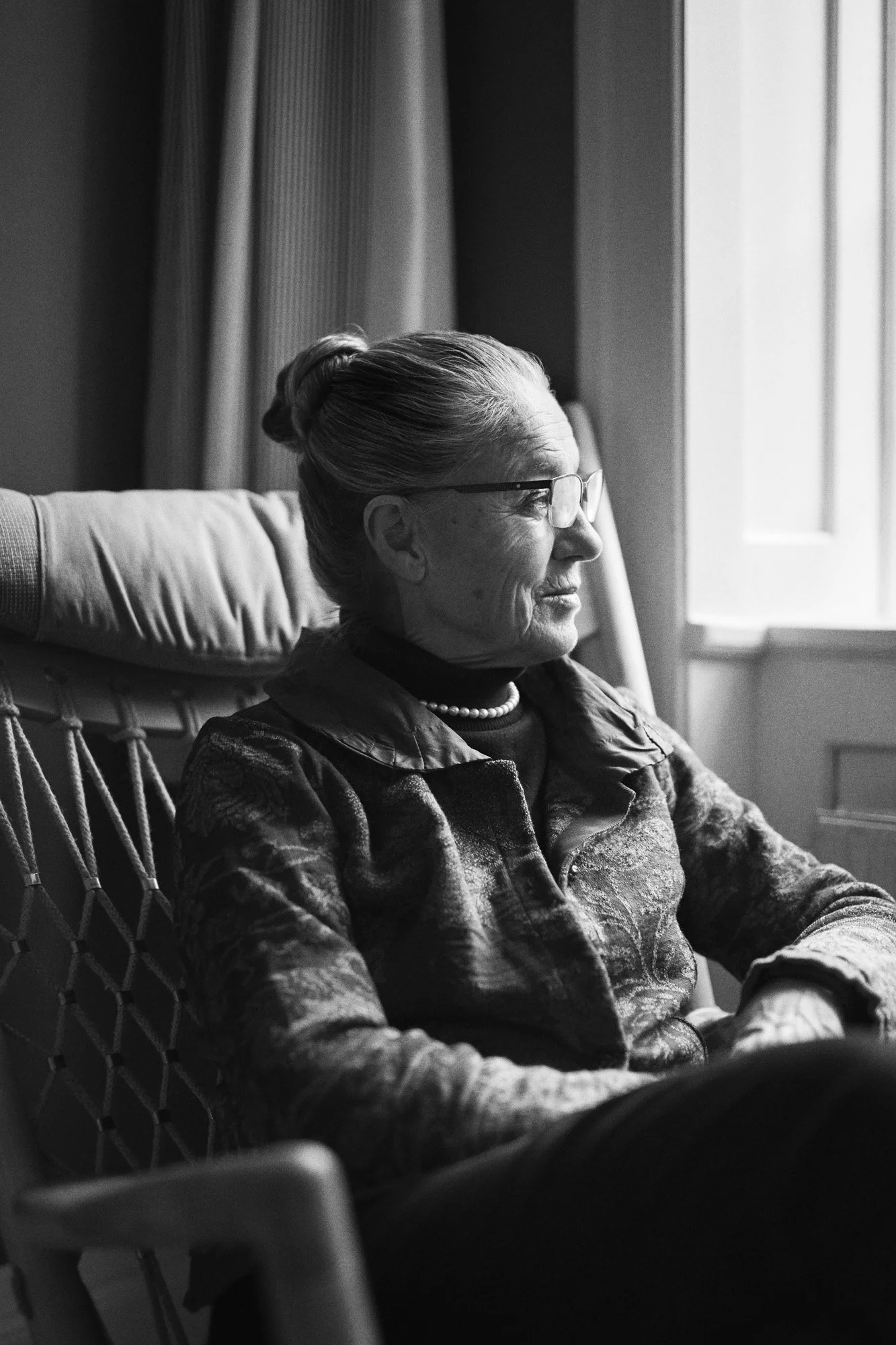 Portrait Ritt Bjerregaard - A black and white photo of an elderly woman sitting by a window, wearing glasses, a pearl necklace, and a patterned jacket, gazing outside thoughtfully.