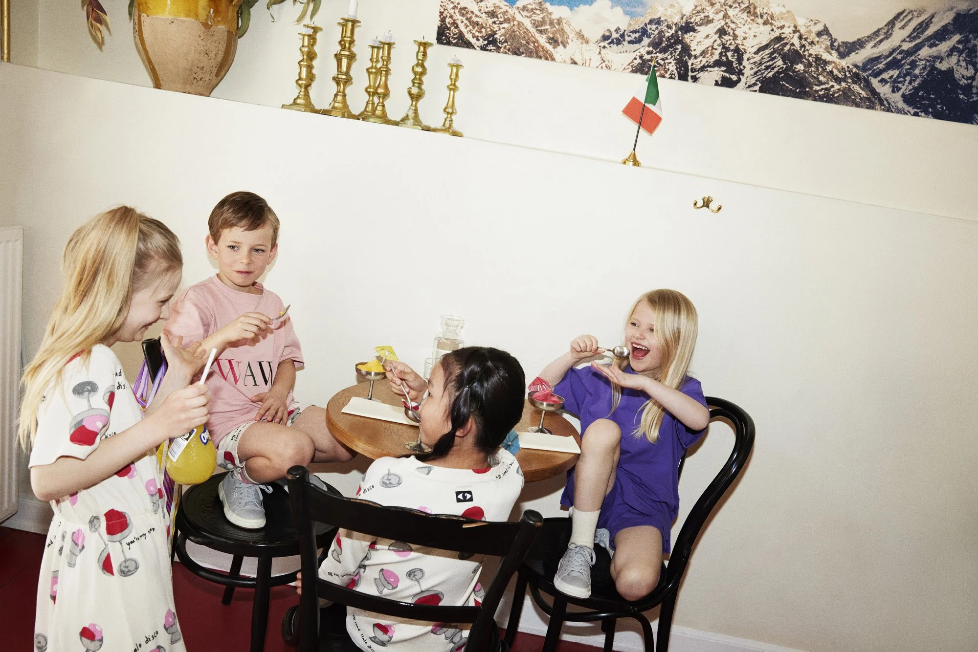 Children celebrating a birthday party with cake and drinks around a table, with a mountain picture and gold candlesticks on the wall in the background.