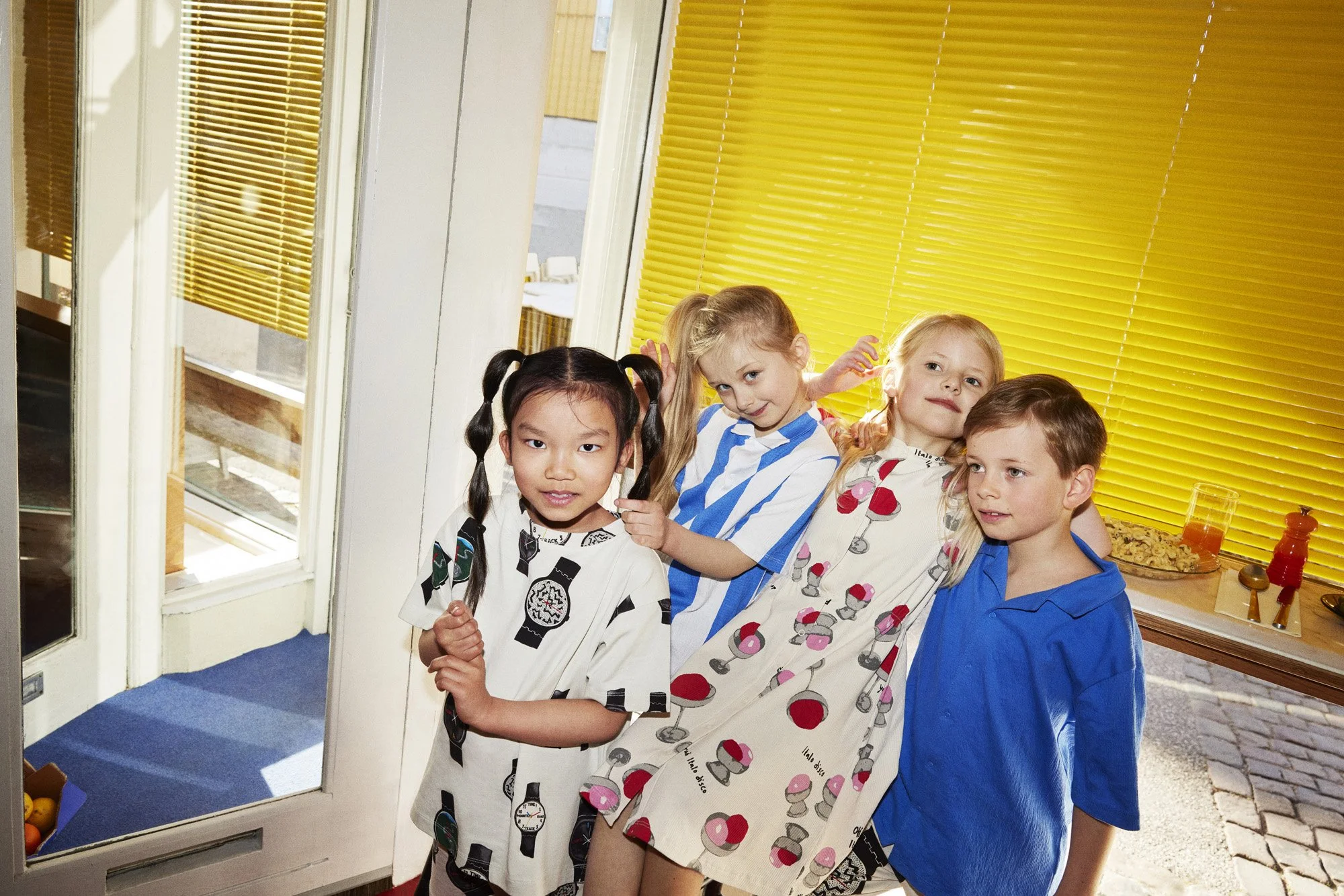 Four children standing indoors near a door with yellow blinds behind them, dressed in colorful casual clothes.
