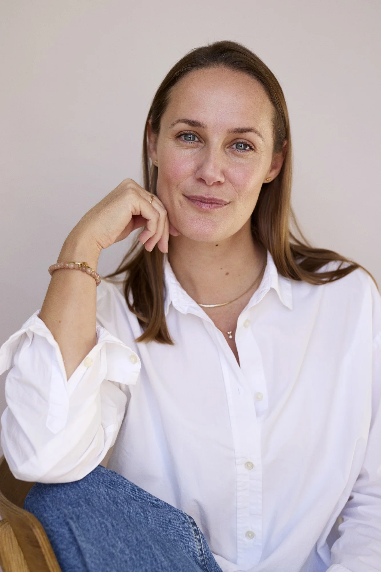 Portrait of a woman with brown hair in a white button-up shirt, sitting with her chin resting on her hand, smiling softly at the camera, against a plain background.