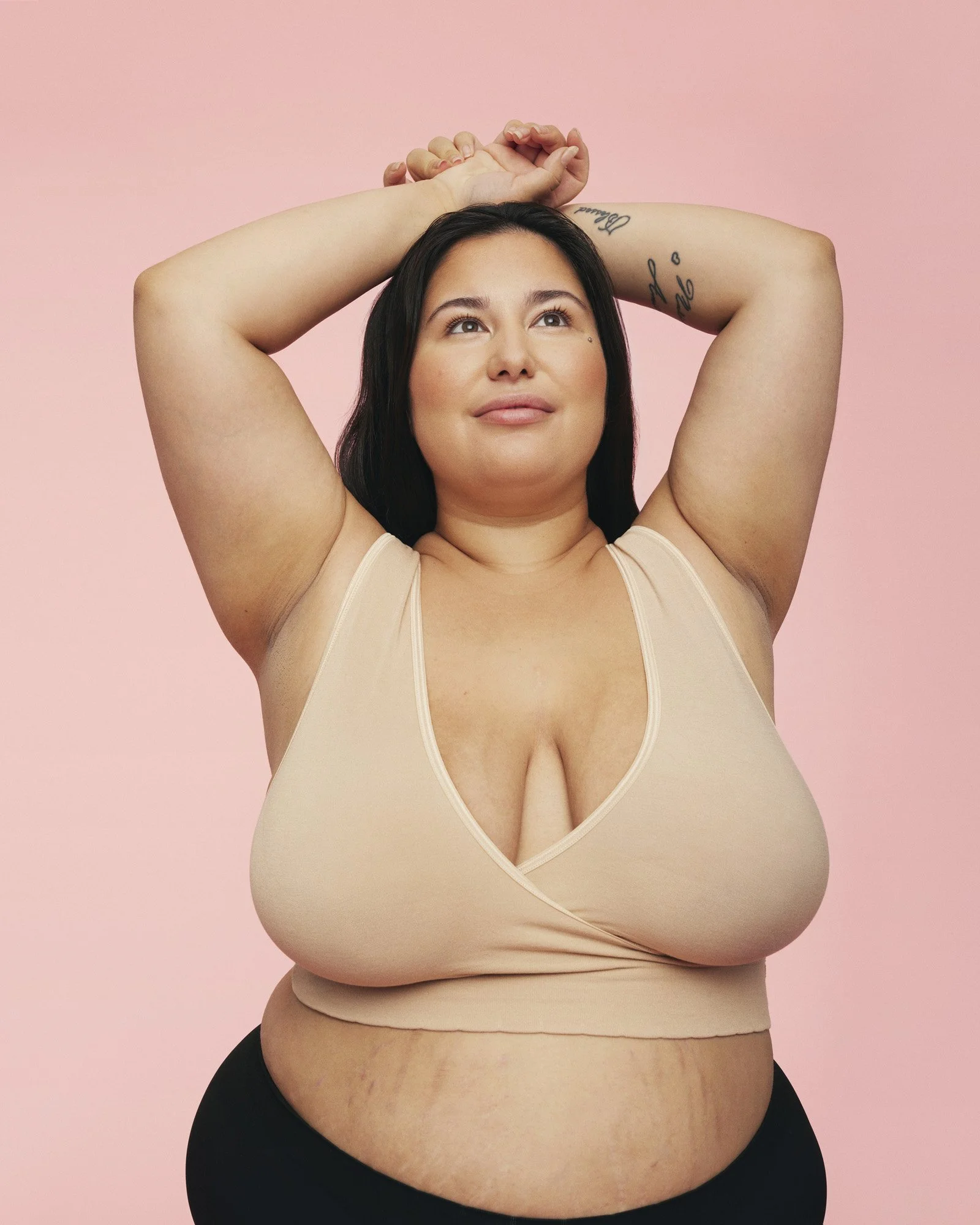MOMKIND - A woman with dark hair and tattoos on her arms, standing with her arms raised above her head against a pink background. She is wearing a beige sleeveless top and black pants, looking slightly upward with a serene expression.