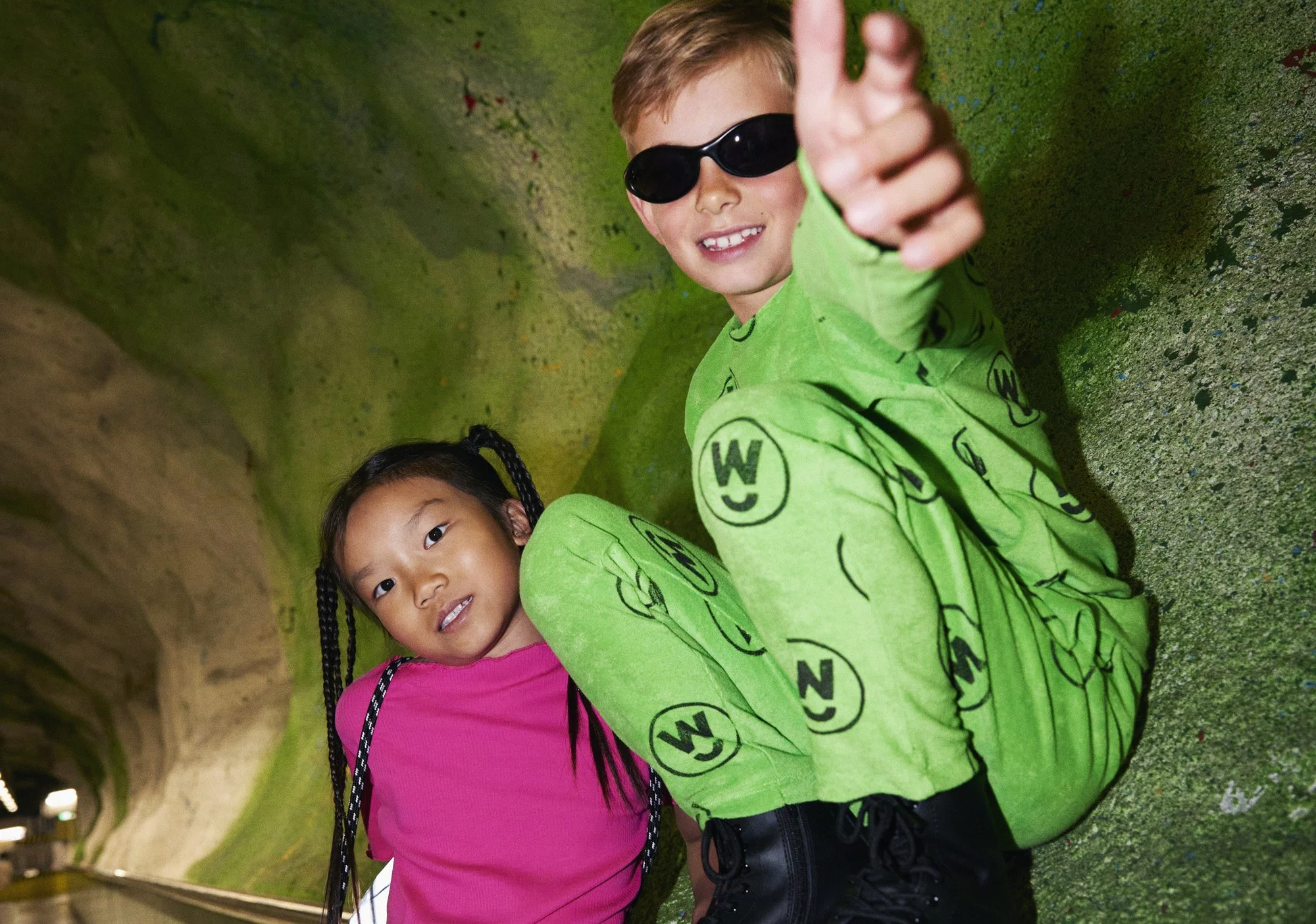 A young boy in a green jumpsuit with black pattern and black sunglasses giving a thumbs-up, and a young girl in a pink shirt with black braids looking at the camera, inside a green, moss-covered tunnel.