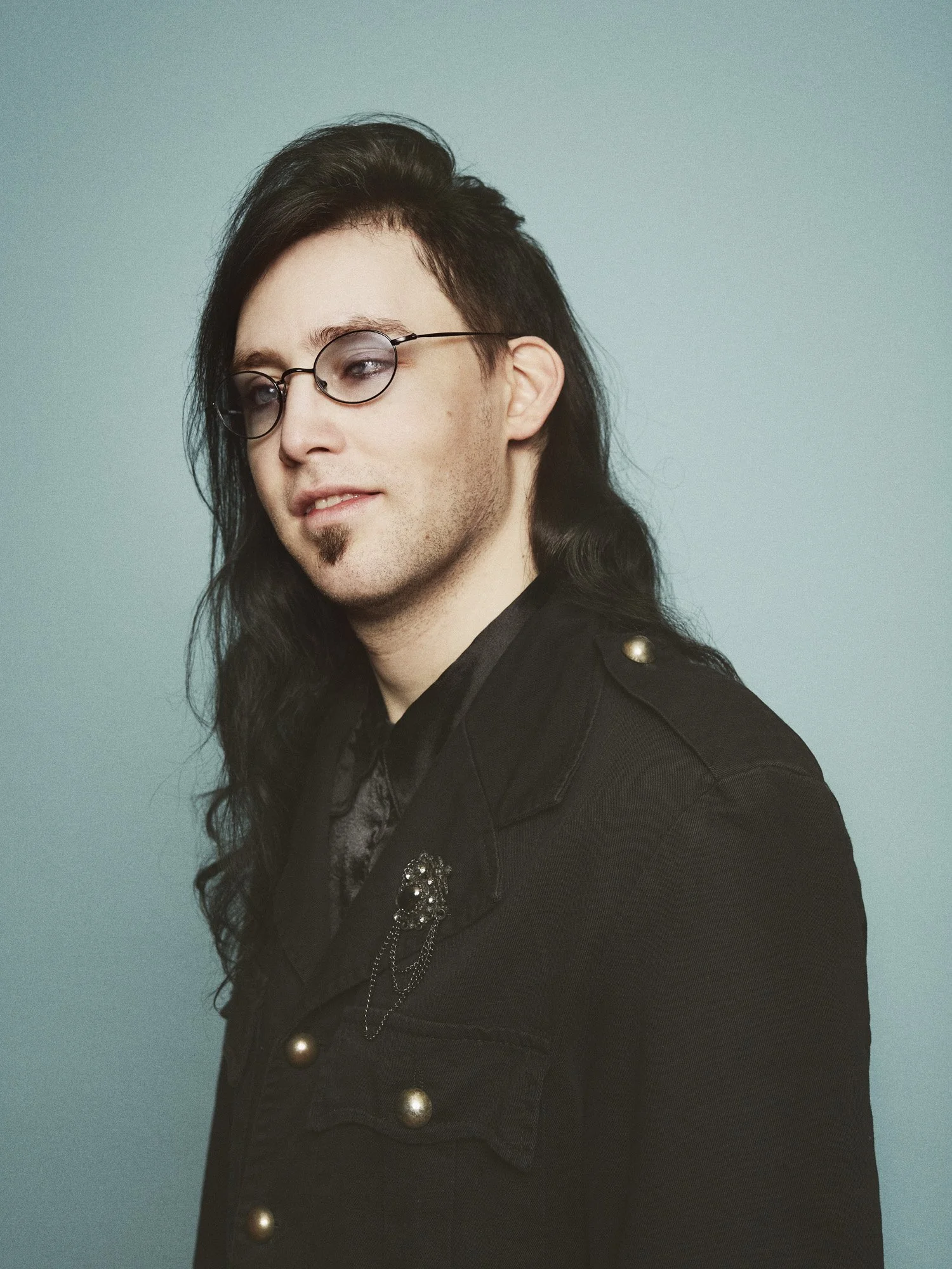GLAD Teater - Portrait of a young man with long black hair, glasses, and a goatee, wearing a black jacket with gold buttons and a brooch, against a light blue background.
