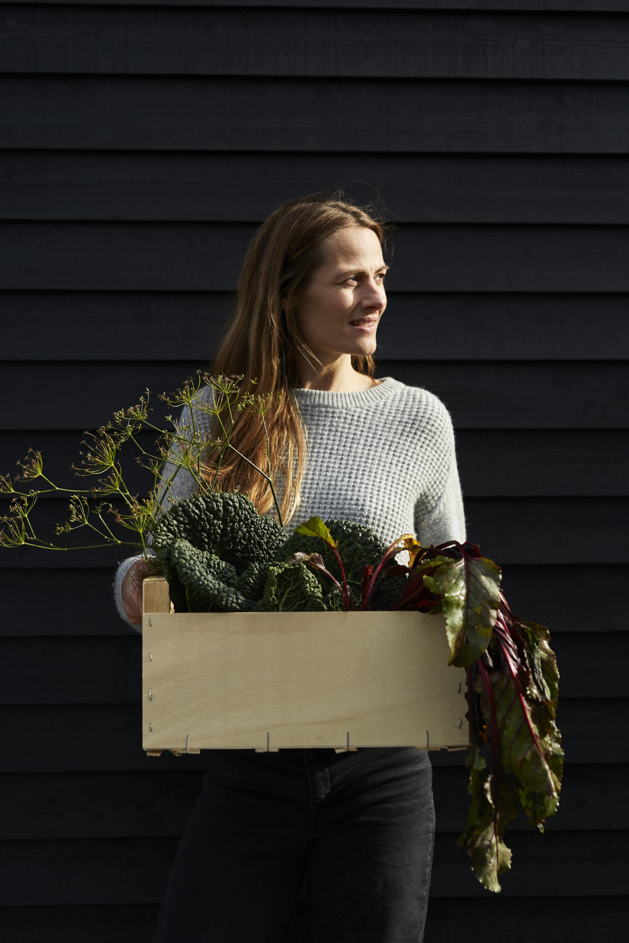 A woman holding a wooden box with leafy vegetables and plants, standing in front of a black wooden wall.