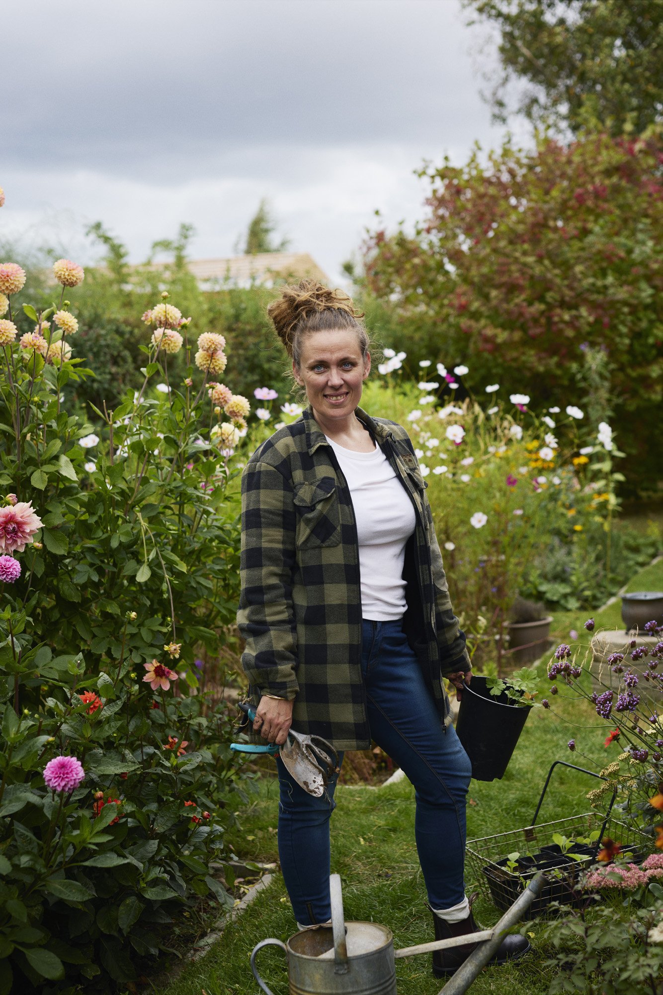 Cristine Glise - Haven Håbets og Kærligheden A woman smiling while standing in a garden, holding a black bucket and gardening tools, surrounded by blooming flowers and plants.