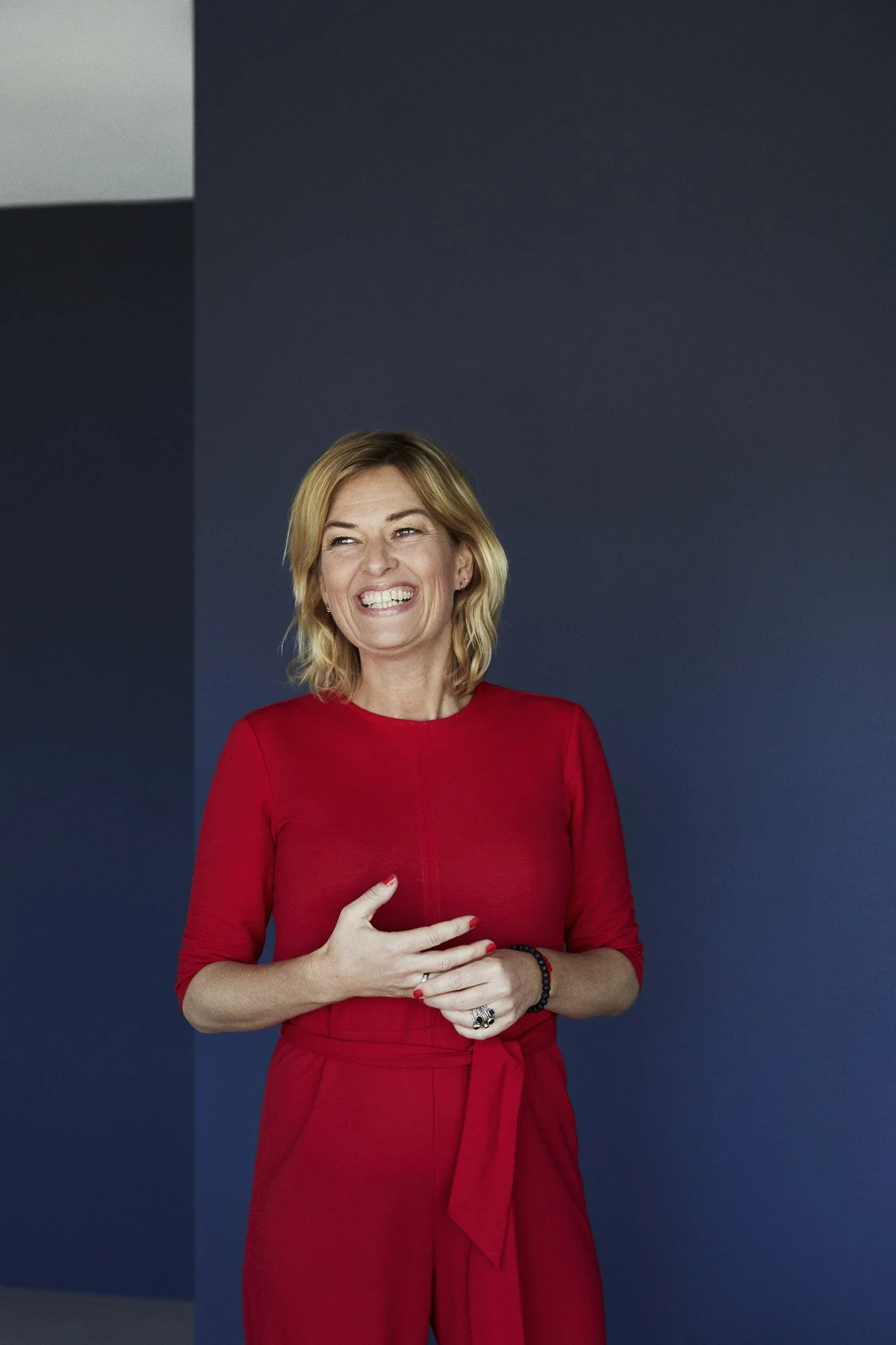 A woman with blonde hair, smiling, wearing a red dress and standing against a dark blue and gray wall.