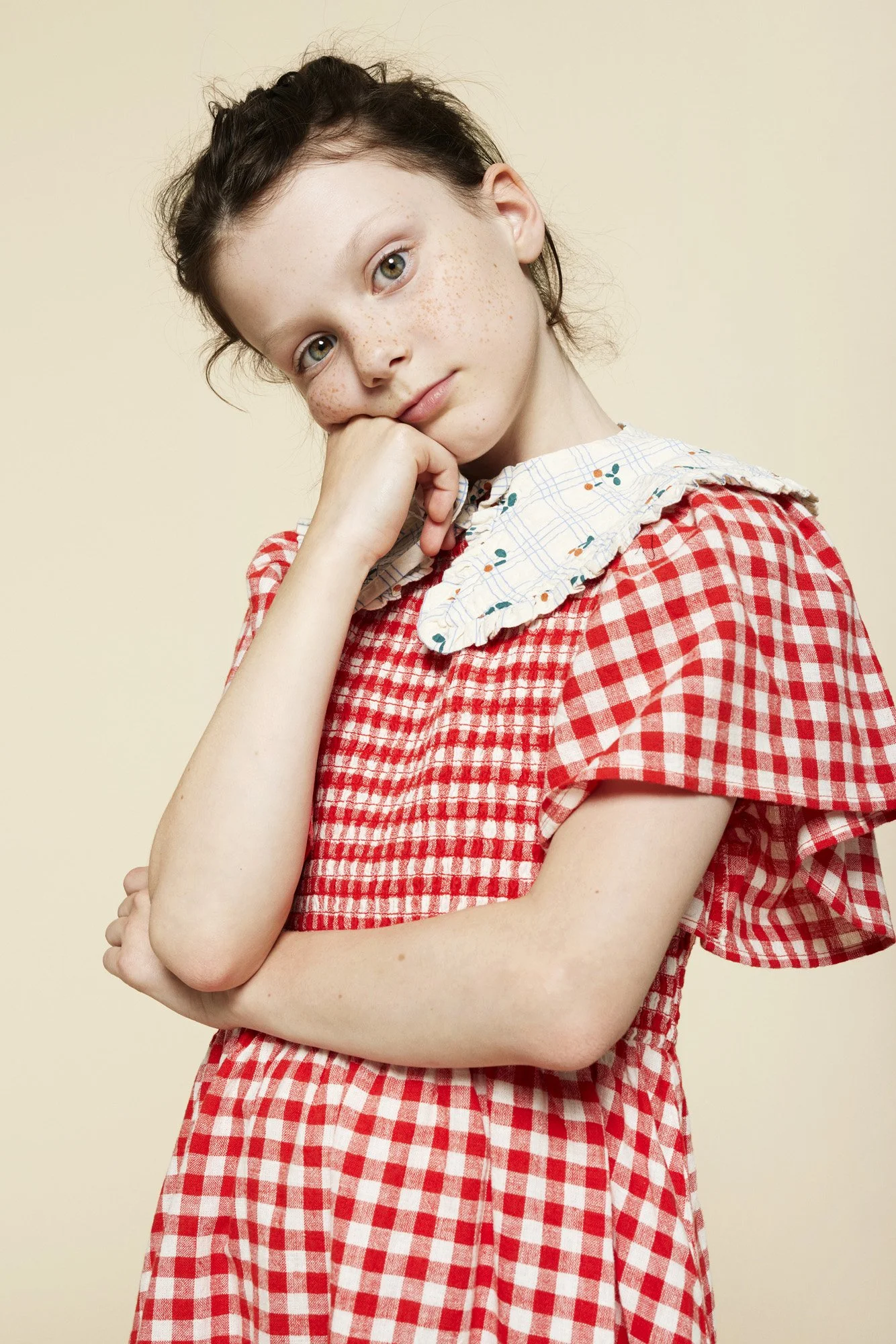 A young girl with light skin and freckles wearing a red and white checkered dress with puffy sleeves and a white ruffled collar with blue, orange, and yellow dots. She has dark brown hair styled with a braid at the top and is posing with her chin res