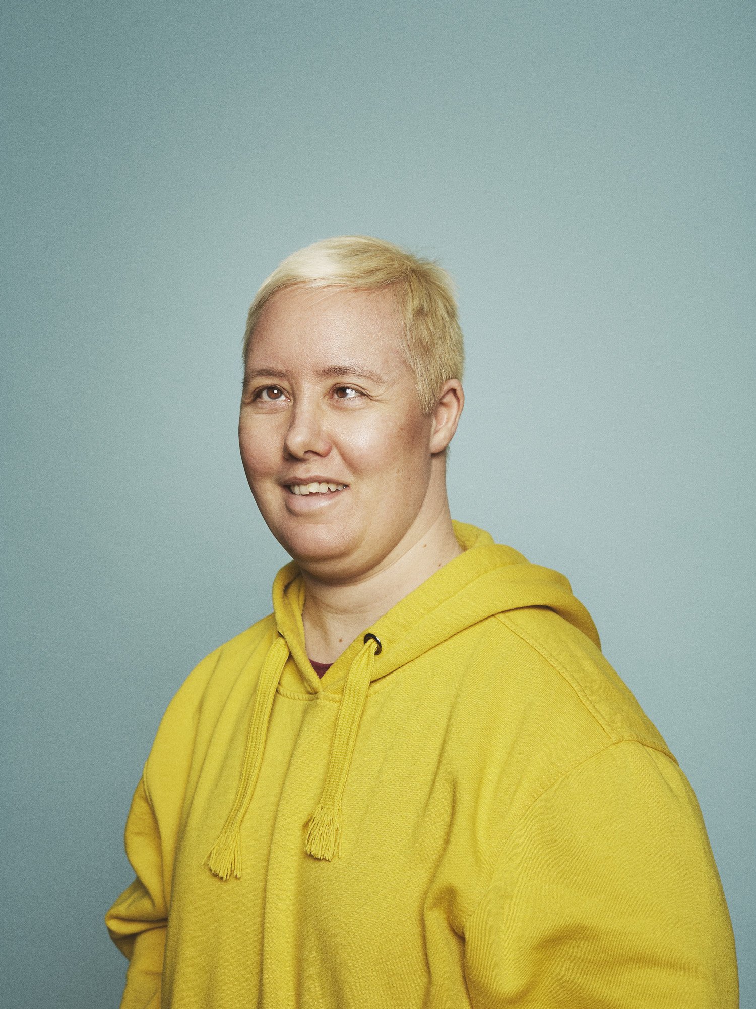 GLAD Teater - Portrait of a person with bleach blonde hair wearing a yellow hoodie against a light blue background.