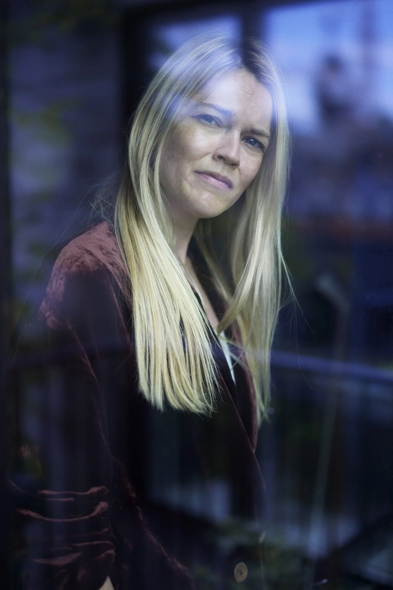 Portrait Psykoterapeut Lena Breum - A woman with long blonde hair and fair skin gazes through a window with a slight smile, wearing a dark reddish-brown jacket.