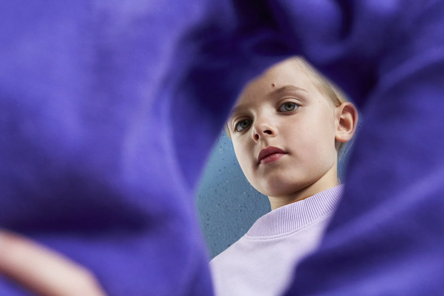 WAWA CPH - A young girl with blond hair and blue eyes looking into a mirror, partially framed by purple clothing or fabric.