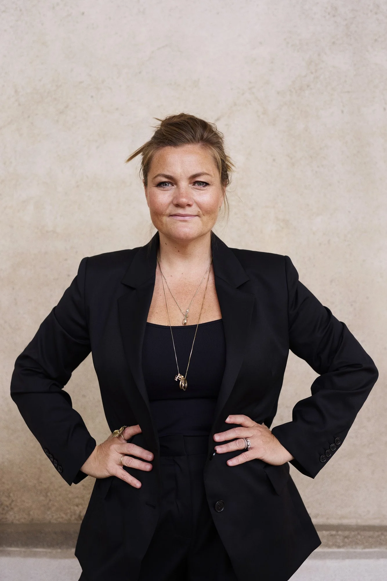 Portrait Thea from Unconventional Ventures - A woman with blonde hair tied back, wearing a black blazer and black top, standing with hands on hips against a beige wall.
