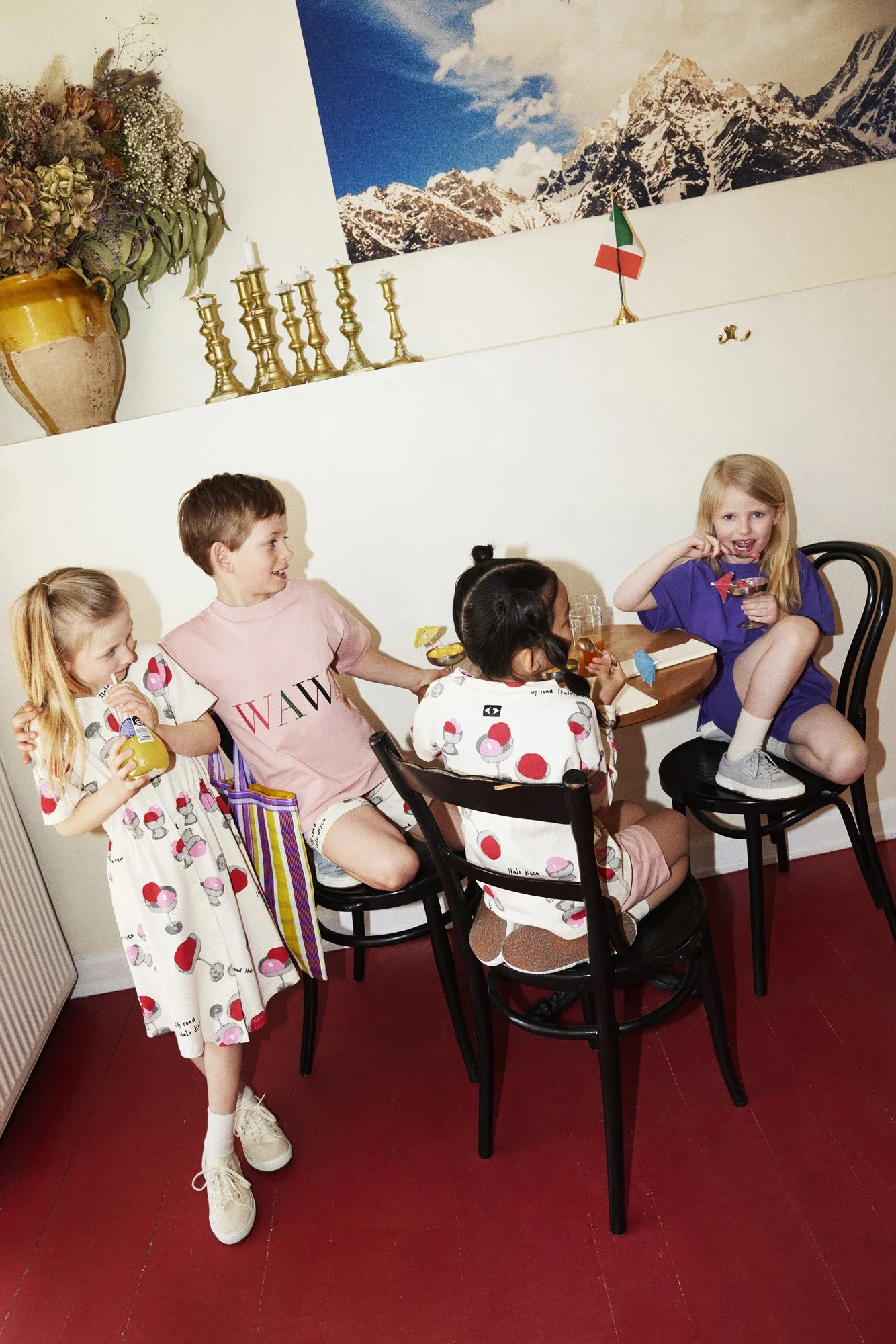 Five children celebrating a birthday party indoors. Two girls and a boy are sitting on chairs, and two girls are standing, enjoying drinks and snacks. The scene includes colorful drinks with umbrellas, a large vase of flowers, a mountain landscape ph